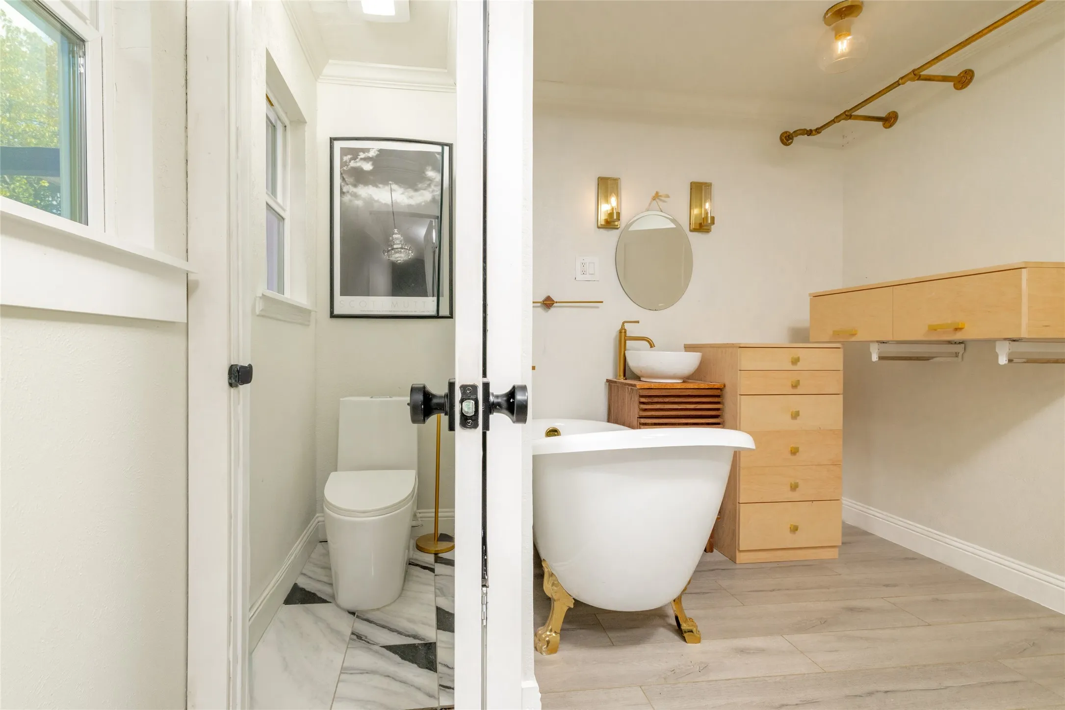 Freestanding tub with gold-finish feet, vessel sink with gold-finish faucet, light wood-finish cabinetry, and gold-finish wall sconces