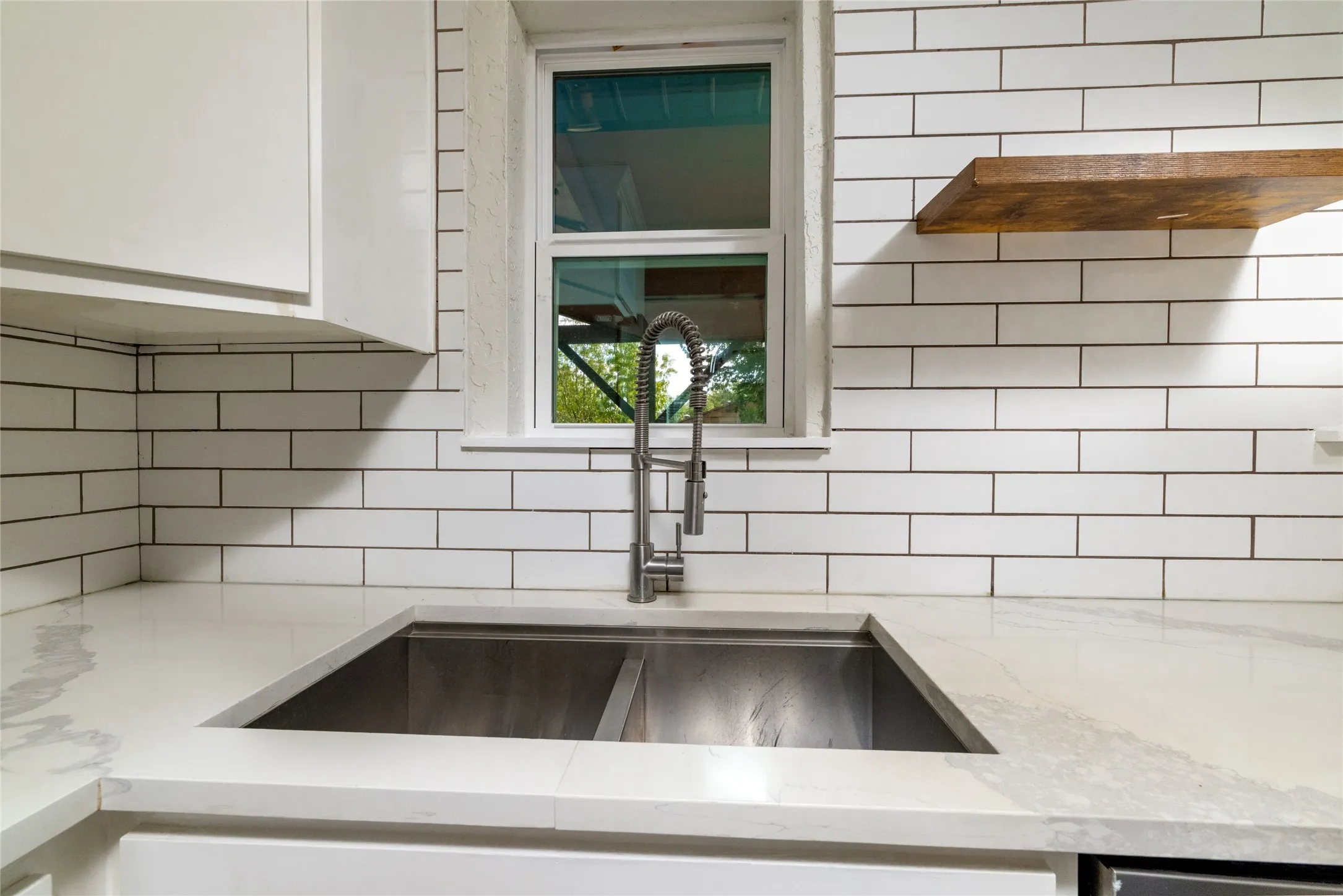Stainless steel double basin sink, gooseneck faucet, white subway tile backsplash, and a floating wood shelf
