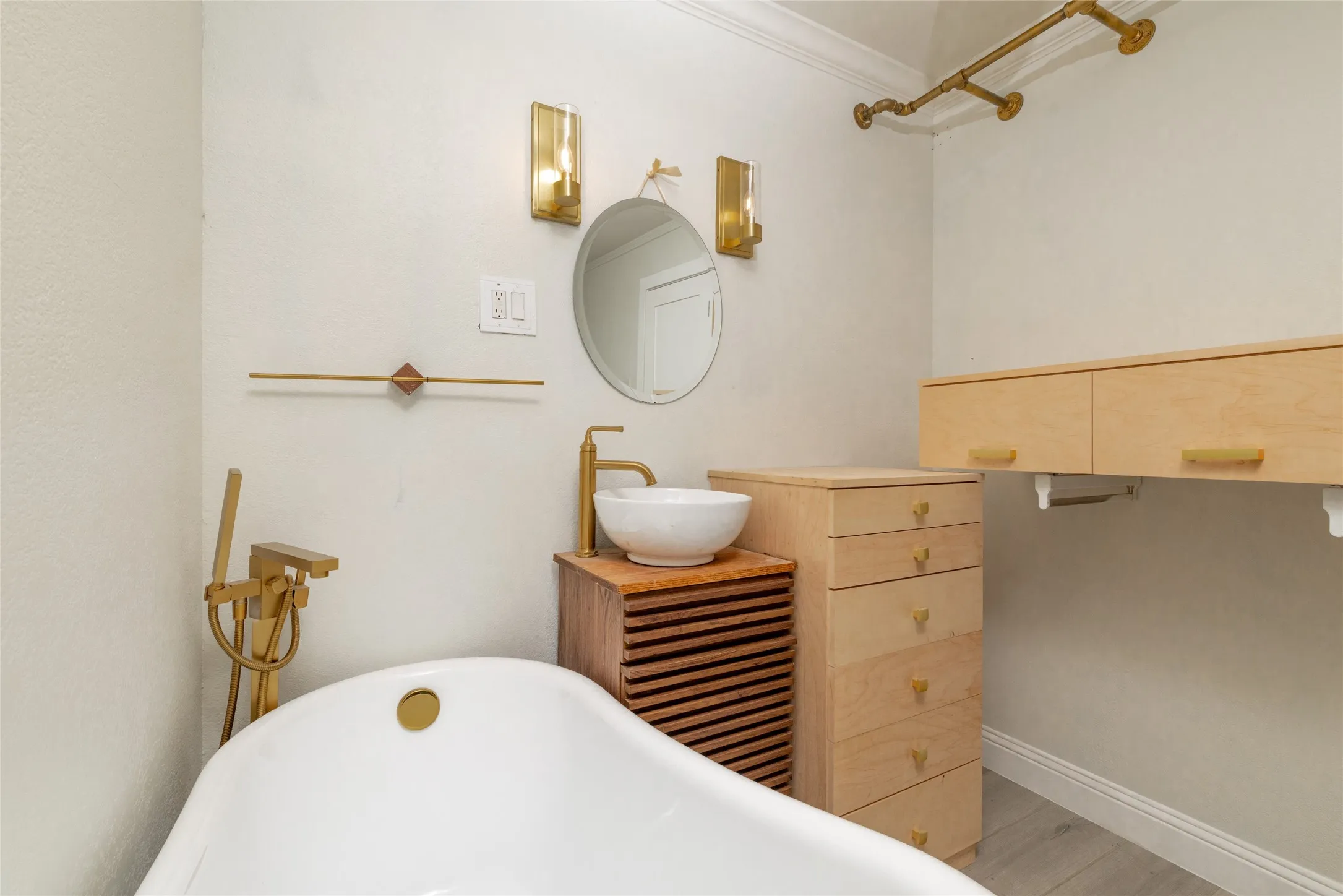 Freestanding soaking tub with floor-mounted brass faucet, vessel sink with brass fixtures, round wall mirror, and modern wall sconces