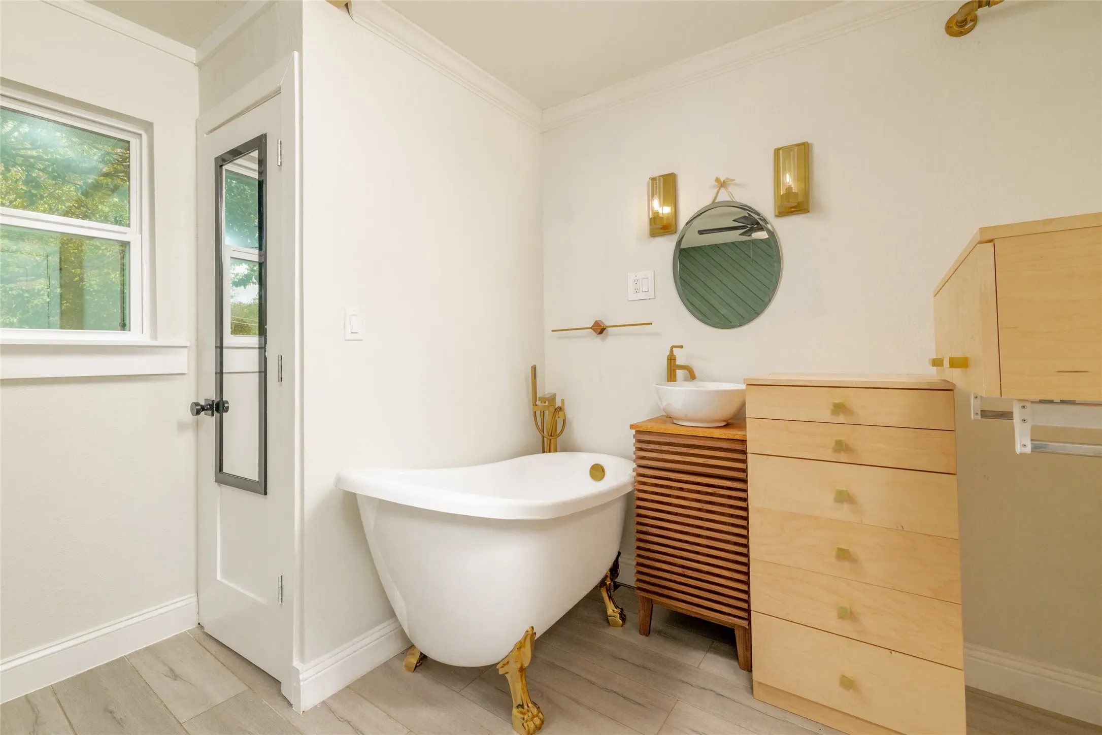 Freestanding soaking tub with ornate gold-finish feet, a white vessel sink on a wood-finish vanity, and light-tone wood-finish built-in storage