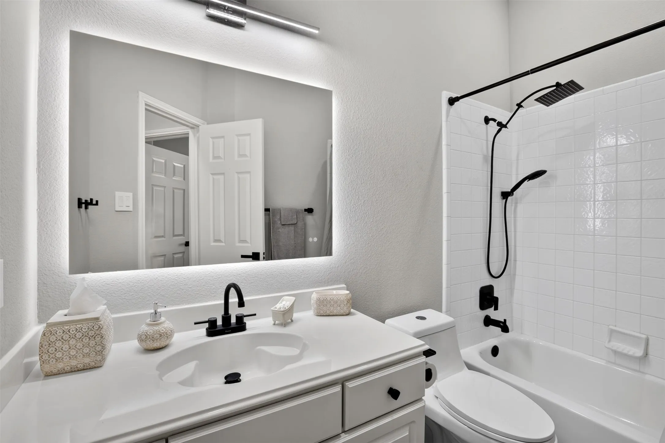 White vanity with an integrated sink and matte black faucet, illuminated mirror with built-in lighting, white subway tile shower surround, and a matte black shower fixture with a handheld wand