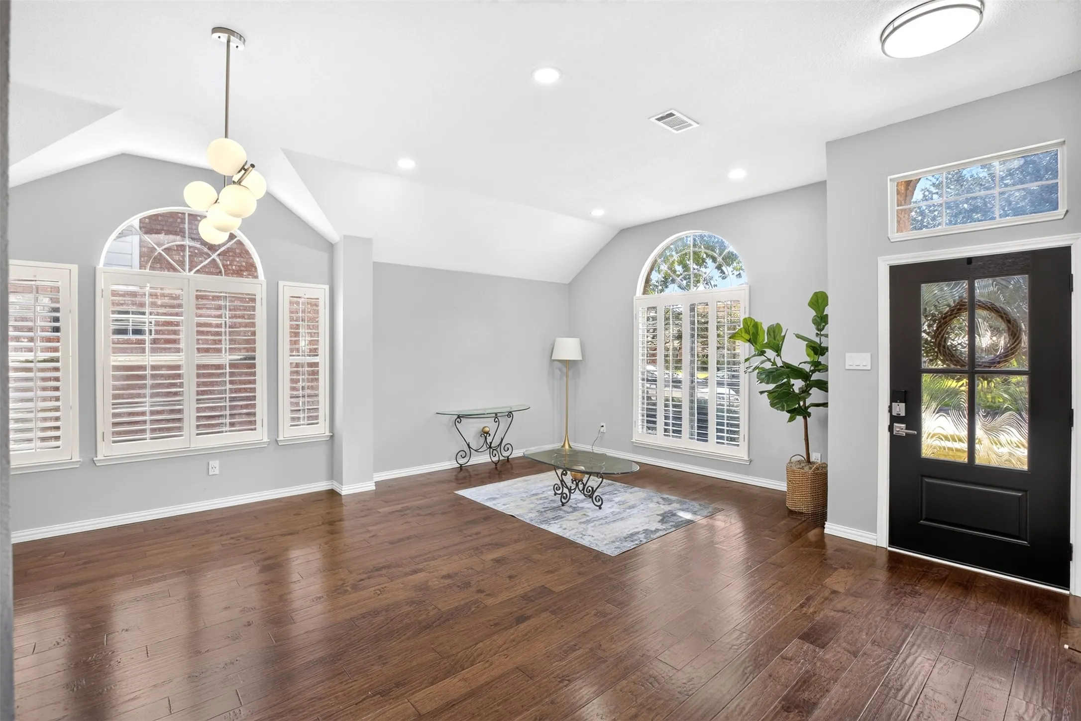 Hardwood-style flooring throughout, vaulted ceilings, arched windows with plantation shutters, recessed lighting, and a black paneled front door with decorative glass inserts