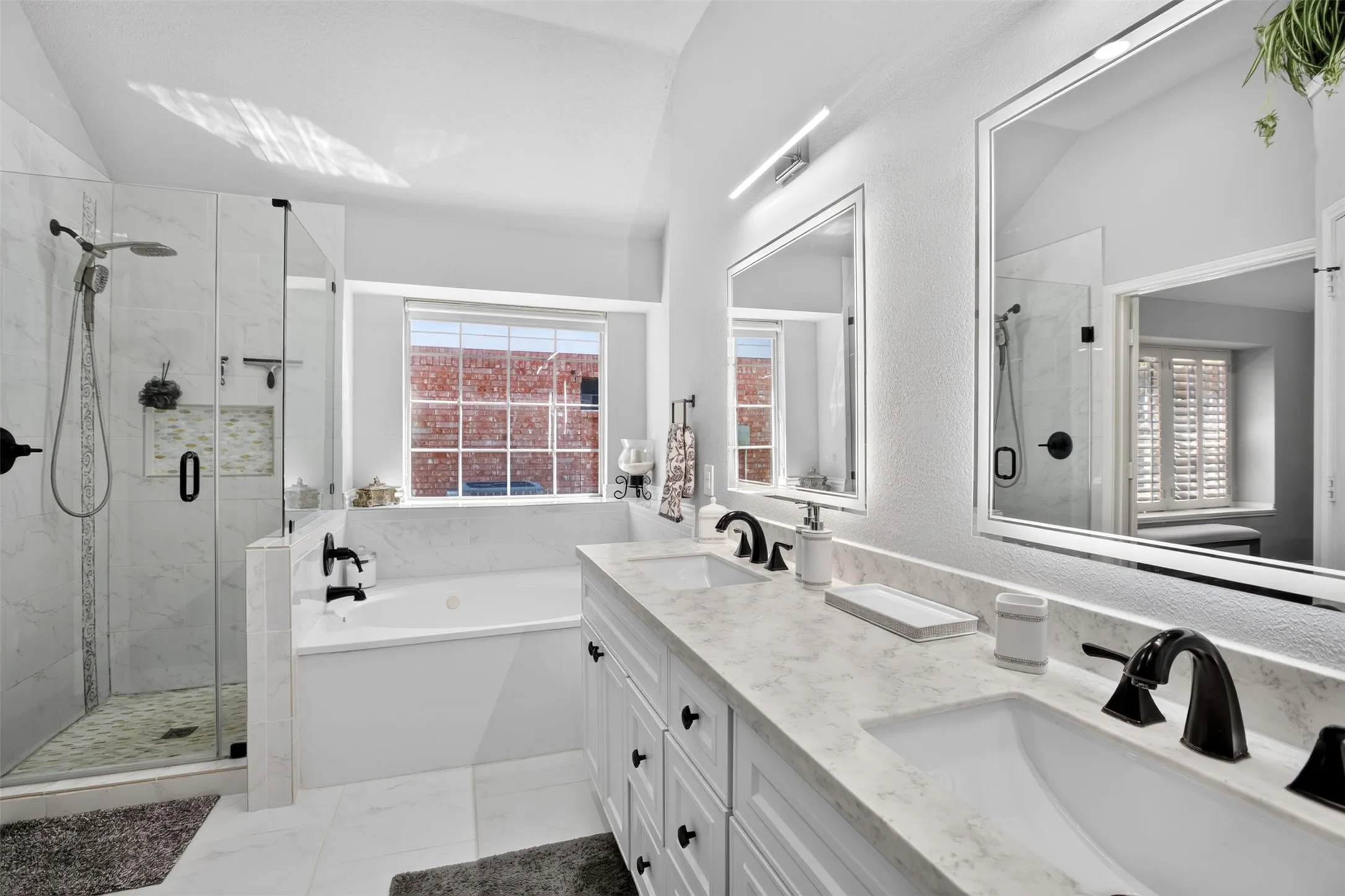 Dual vanity with integrated sinks and black fixtures, frameless glass shower enclosure, built-in soaking tub, large wall-mounted mirrors with integrated lighting, and tile flooring