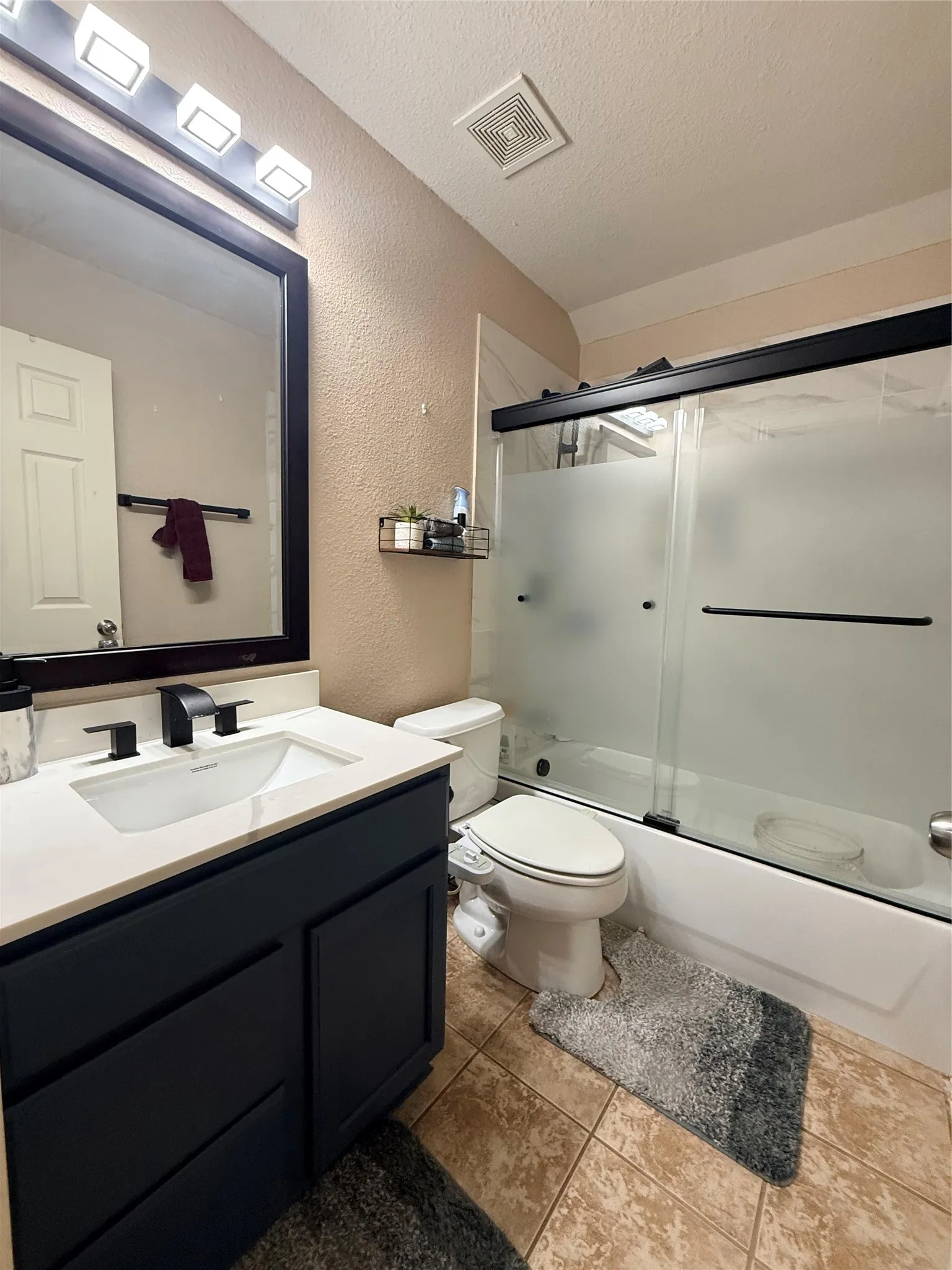 Bathroom featuring a dark vanity with a white countertop and rectangular undermount sink, matte black faucet, framed mirror, and a shower-tub combination with sliding glass doors