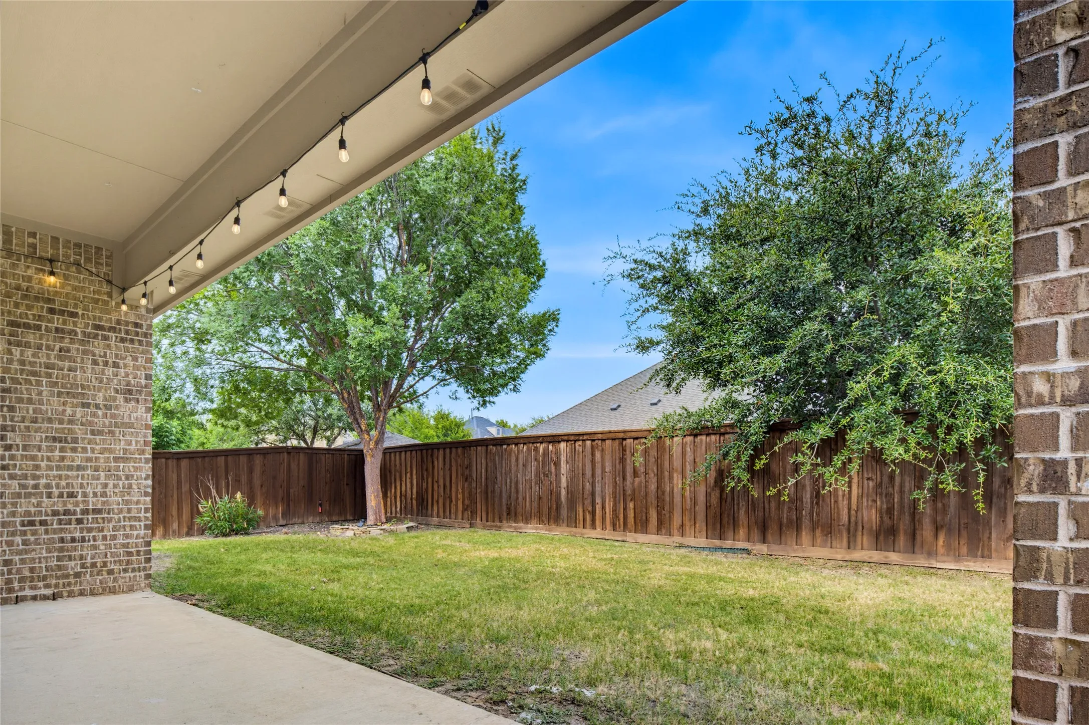 Covered patio with string lighting, brick exterior, concrete patio, wood privacy fence, and mature trees