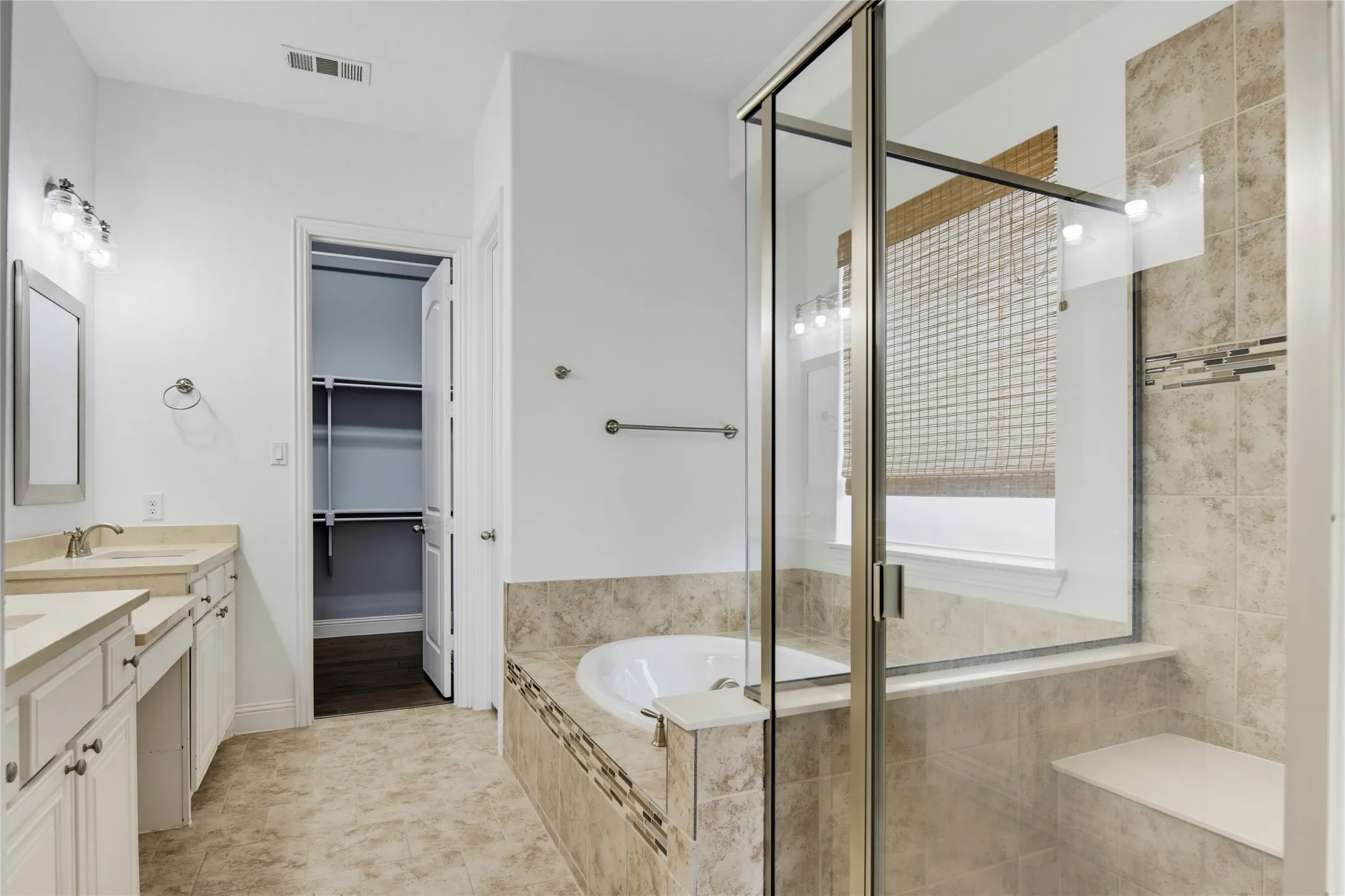 Dual vanity with neutral countertops, built-in soaking tub, glass-enclosed shower with bench, and neutral tile flooring