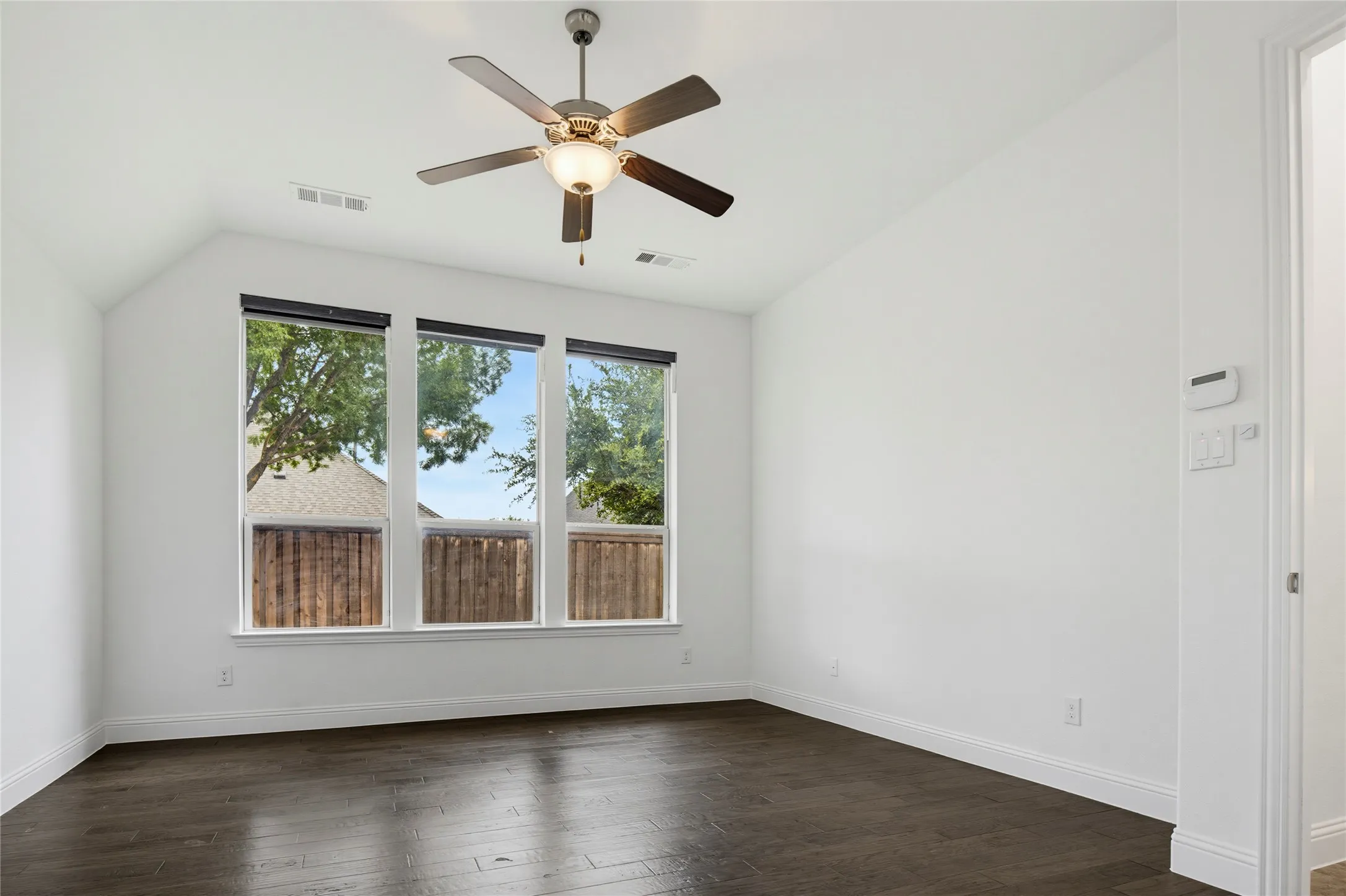 Vaulted ceiling, dark wood-finish flooring, three windows with roller shades, ceiling fan with integrated lighting, and white trim throughout