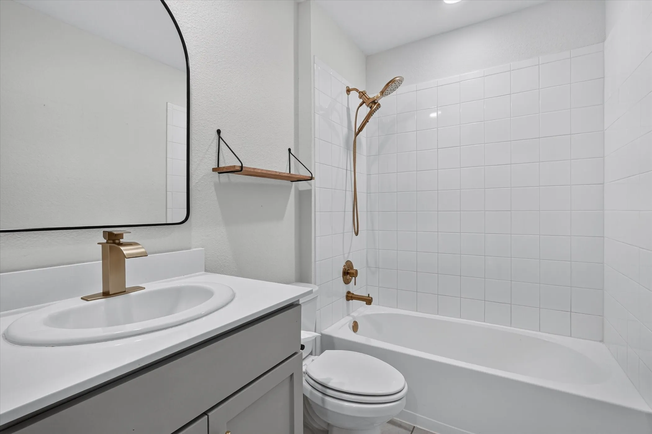 White tile shower-tub combination, single vanity with light gray cabinetry, white countertop, and gold-tone faucet, and an arched mirror