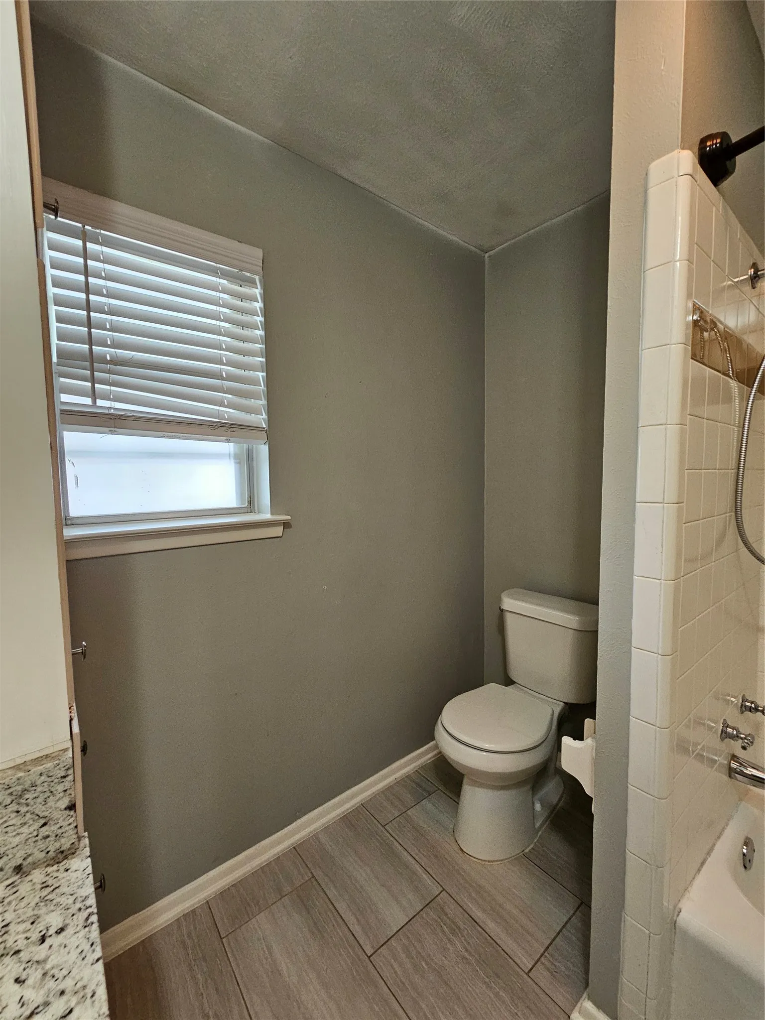 White toilet, light gray tile flooring, window with blinds, and tiled shower surround
