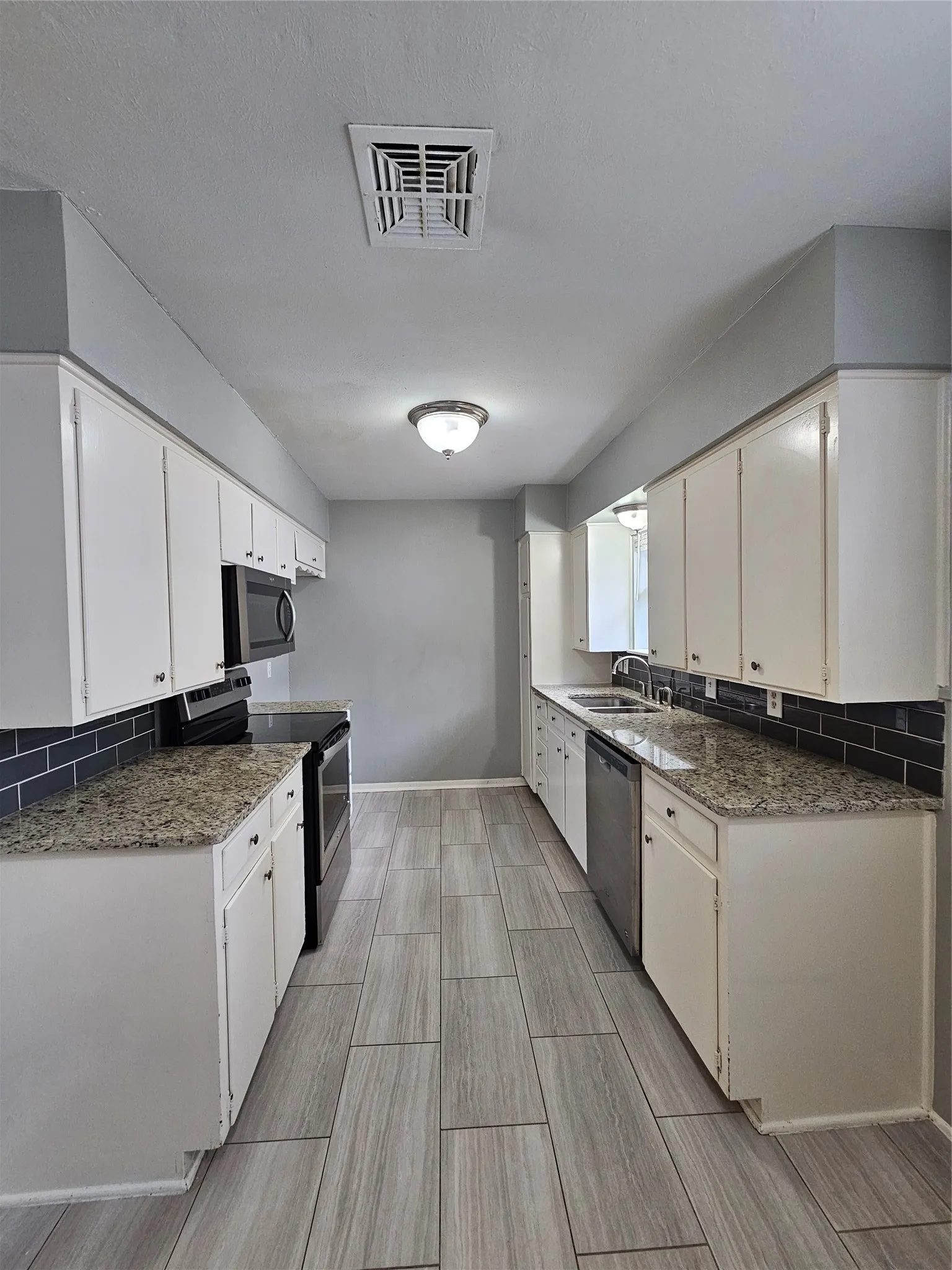 Galley kitchen featuring wood-look tile flooring, white cabinetry, speckled countertops, stainless steel appliances, and a dark subway tile backsplash