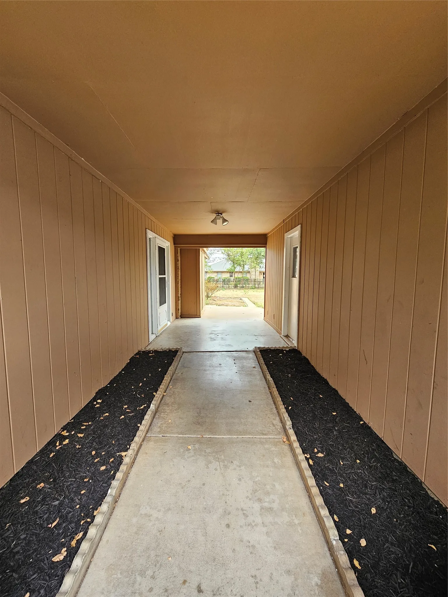 Covered walkway with panel siding, concrete path, and garden beds with dark mulch