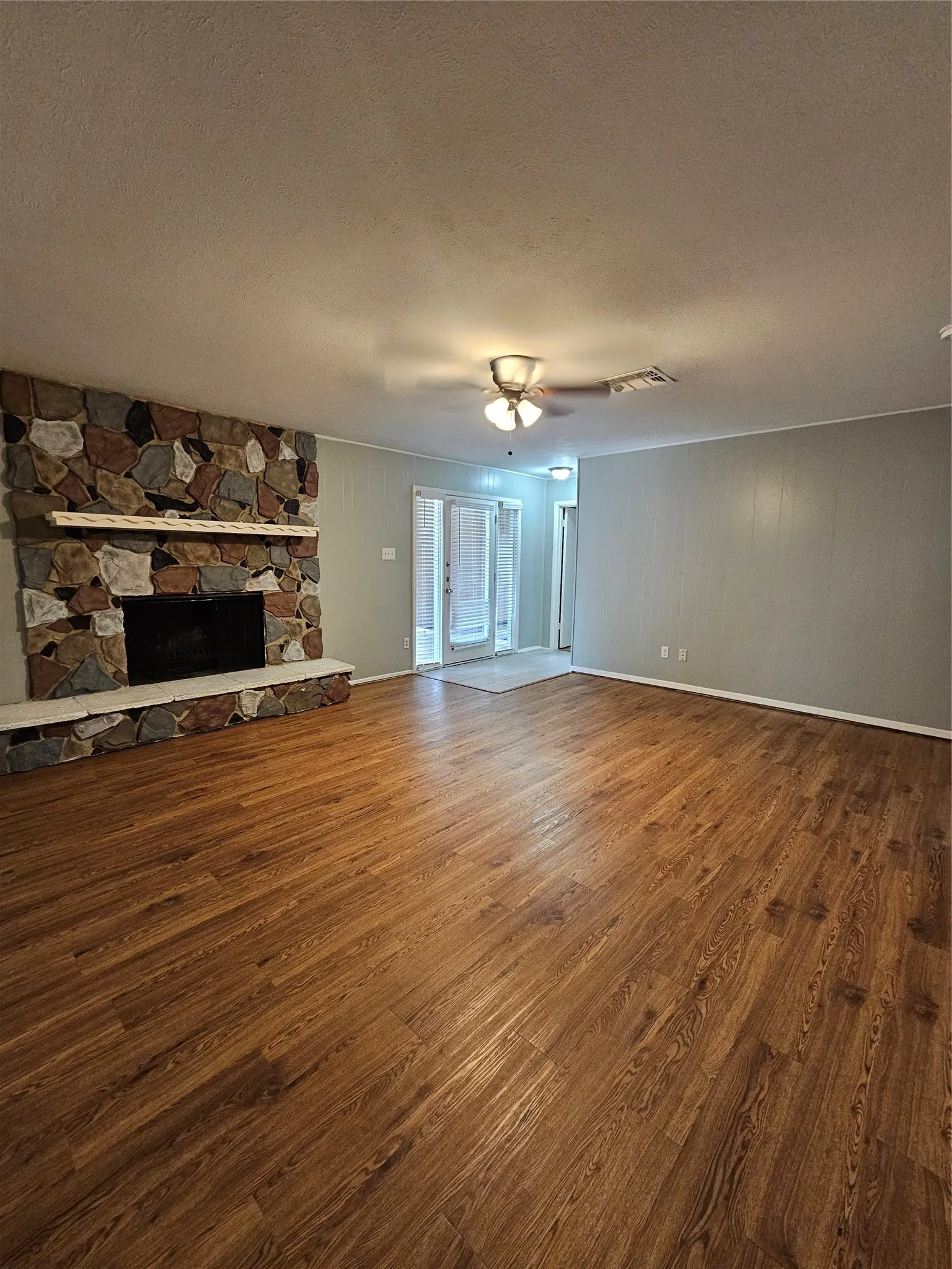 Wood-finish flooring throughout, stone fireplace with a white mantel, ceiling fan with integrated lighting, and double glass doors