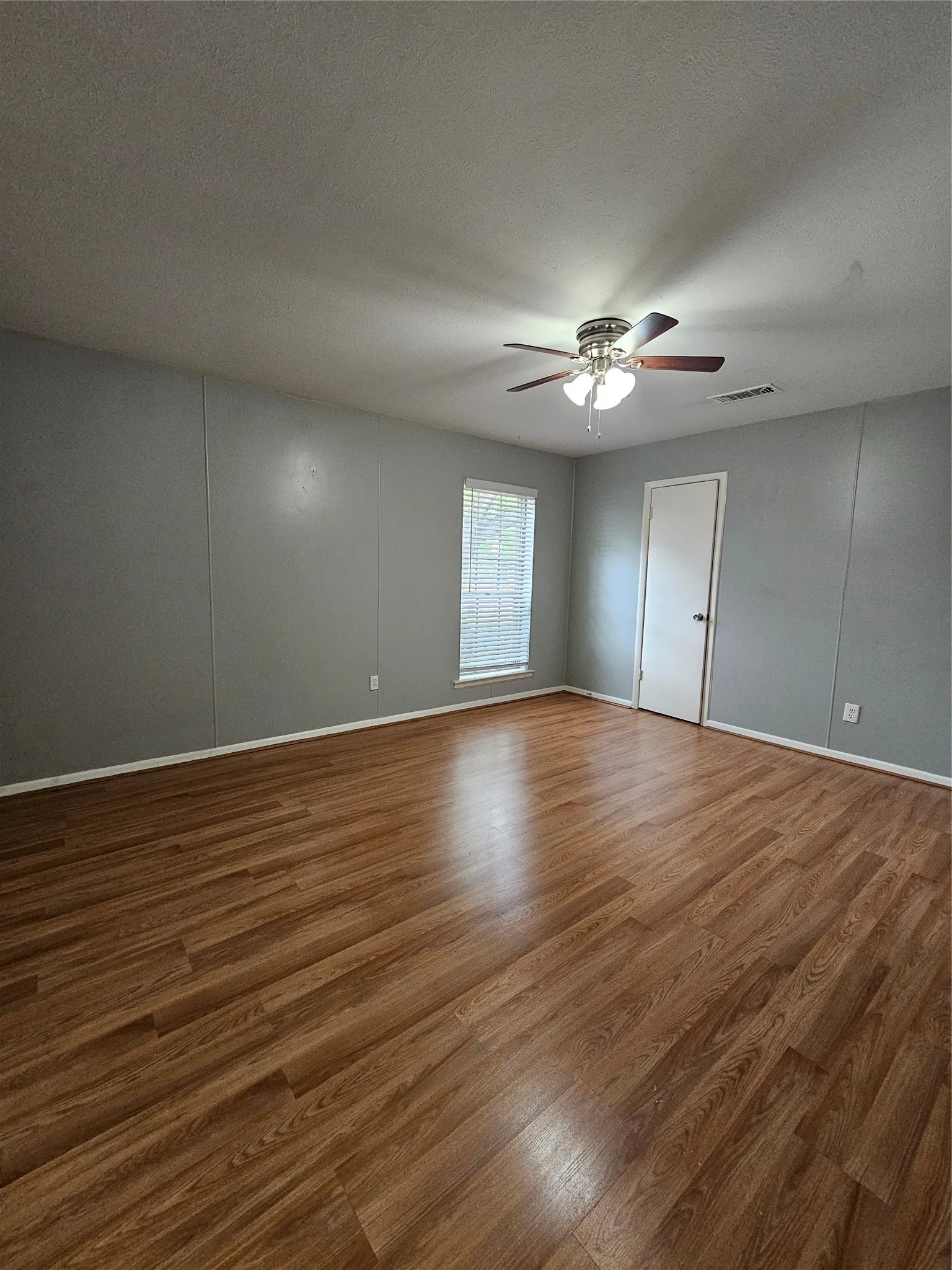 Spacious room featuring wood-finish flooring, light grey walls, a ceiling fan with integrated lighting, and a window with blinds