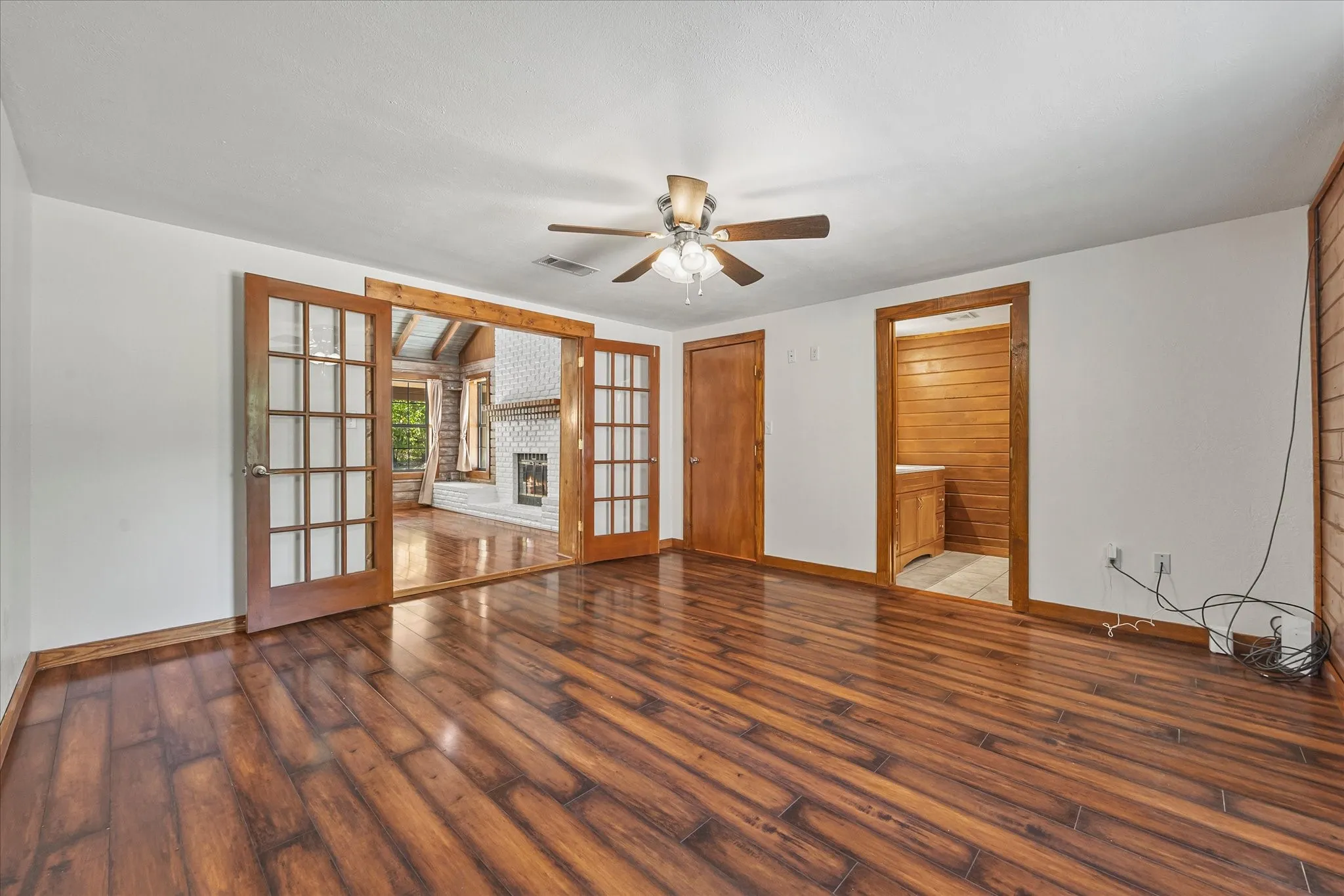 Spacious primary bedroom with ensuite bath, plank floors, french doors leading to living room, log accent wall, and ceiling fan.