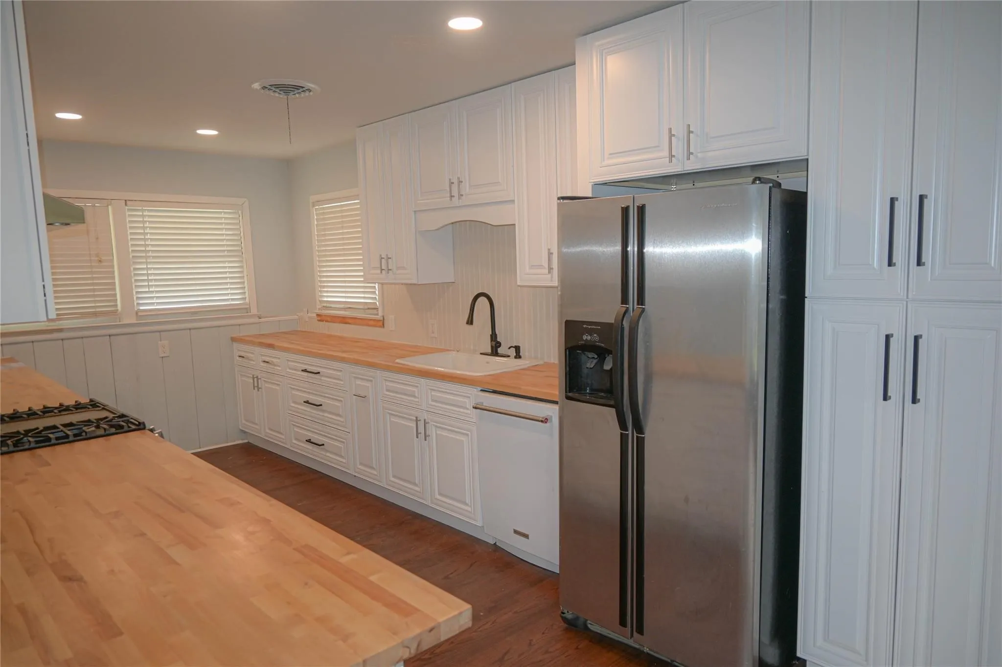 White cabinetry with matte black hardware, wood-finish countertops, stainless steel refrigerator, integrated cooktop, and recessed lighting