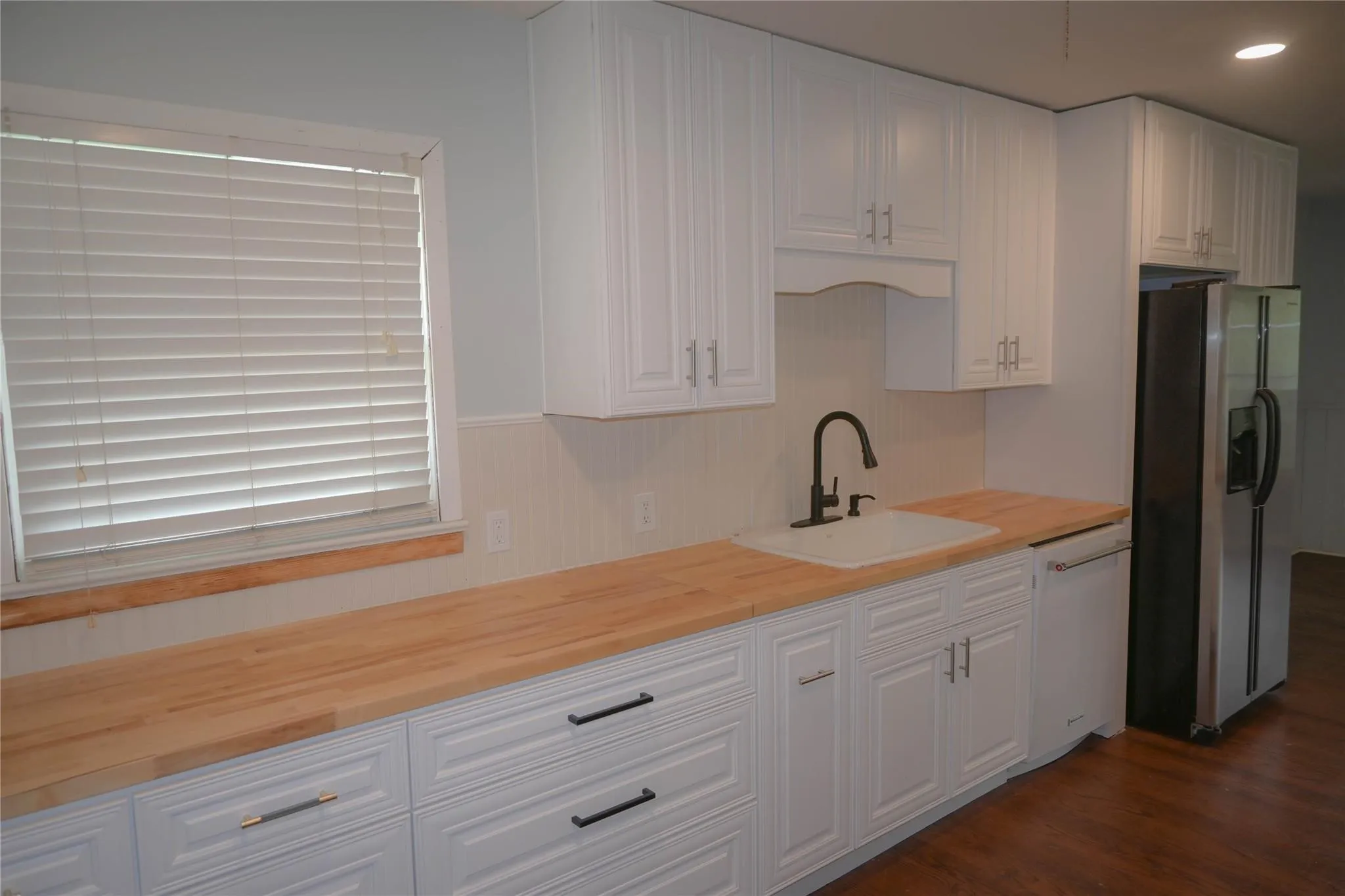 White shaker-style cabinetry with matte black hardware, wood-finish countertops, undermount sink with a matte black faucet, and a stainless steel refrigerator
