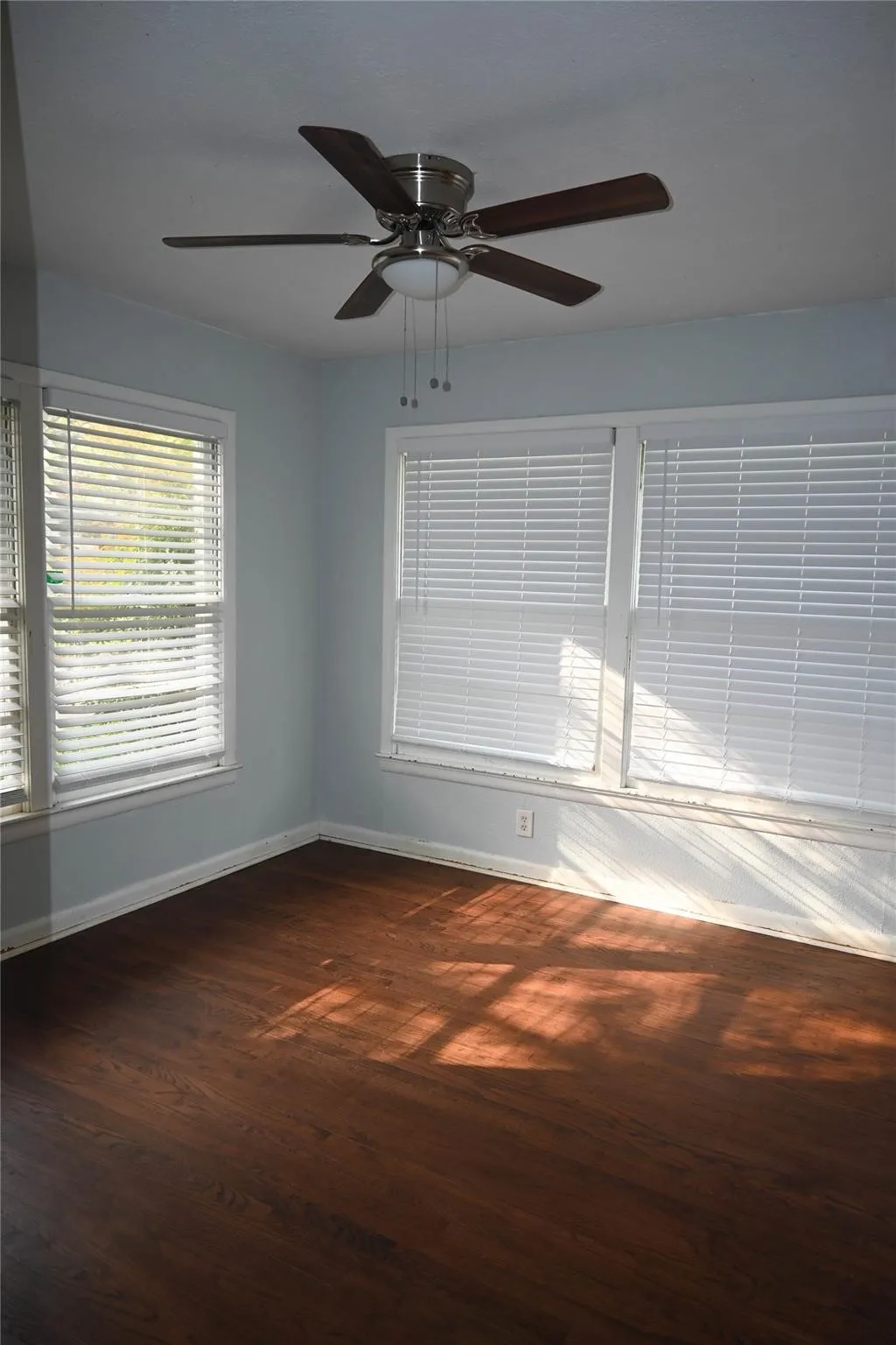 Wood-finish flooring, two large windows with blinds, light blue wall paint, and a ceiling fan with integrated lighting