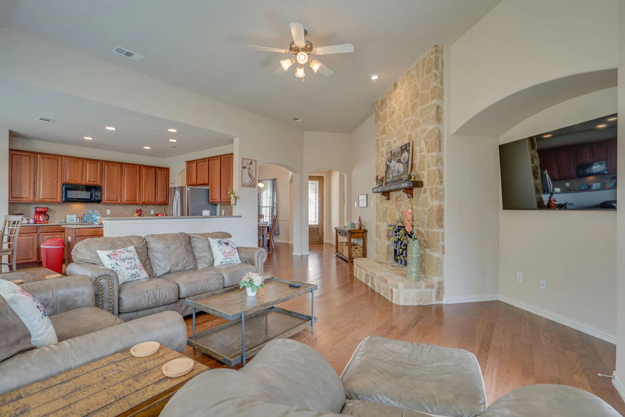 Vaulted ceiling with a ceiling fan, stone-clad fireplace with a wood mantle, wood-finish flooring, recessed lighting, and a kitchen featuring wood cabinetry