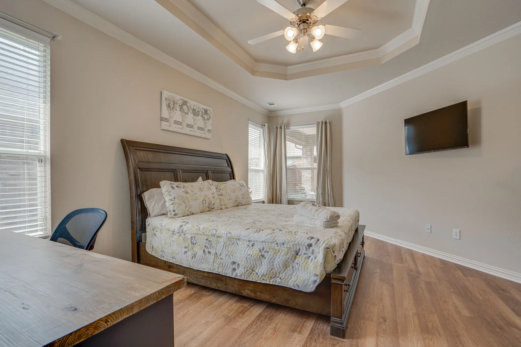 Tray ceiling with a ceiling fan, wall-mounted television, wood-finish flooring, and multiple windows with blinds