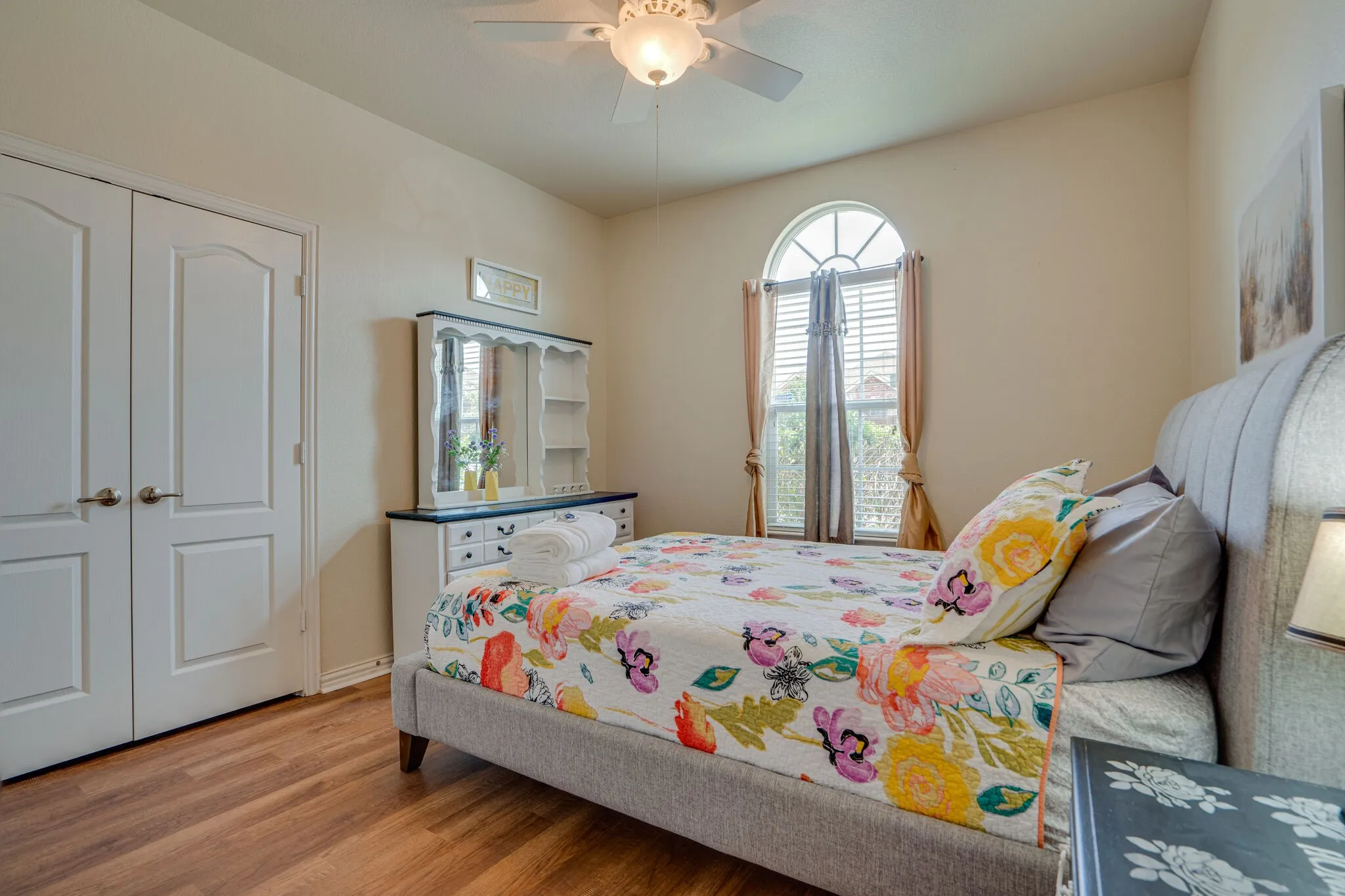Wood-finish flooring, white double closet doors, arched window with blinds, ceiling fan with light fixture