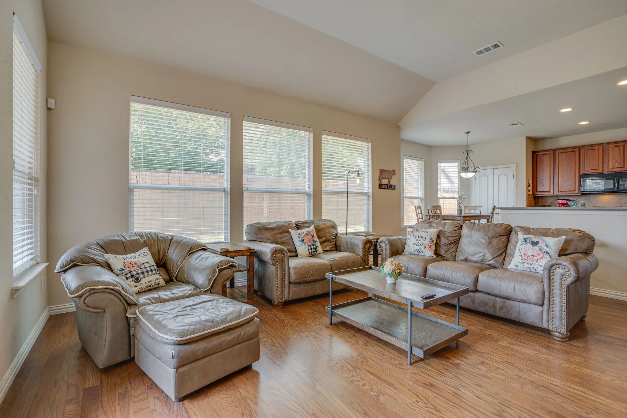 Wood-finish flooring, multiple windows with blinds, vaulted ceiling, recessed lighting, and a partial view of kitchen cabinetry