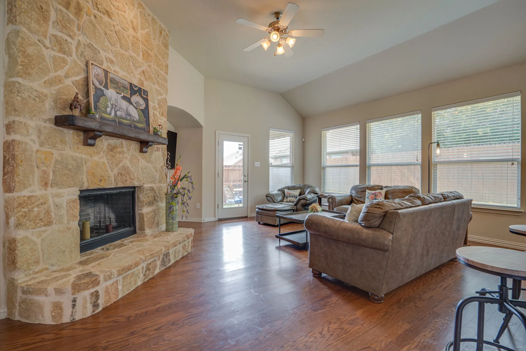 Stone-clad fireplace with mantel, vaulted ceilings, wood-finish flooring, multiple large windows, and a ceiling fan