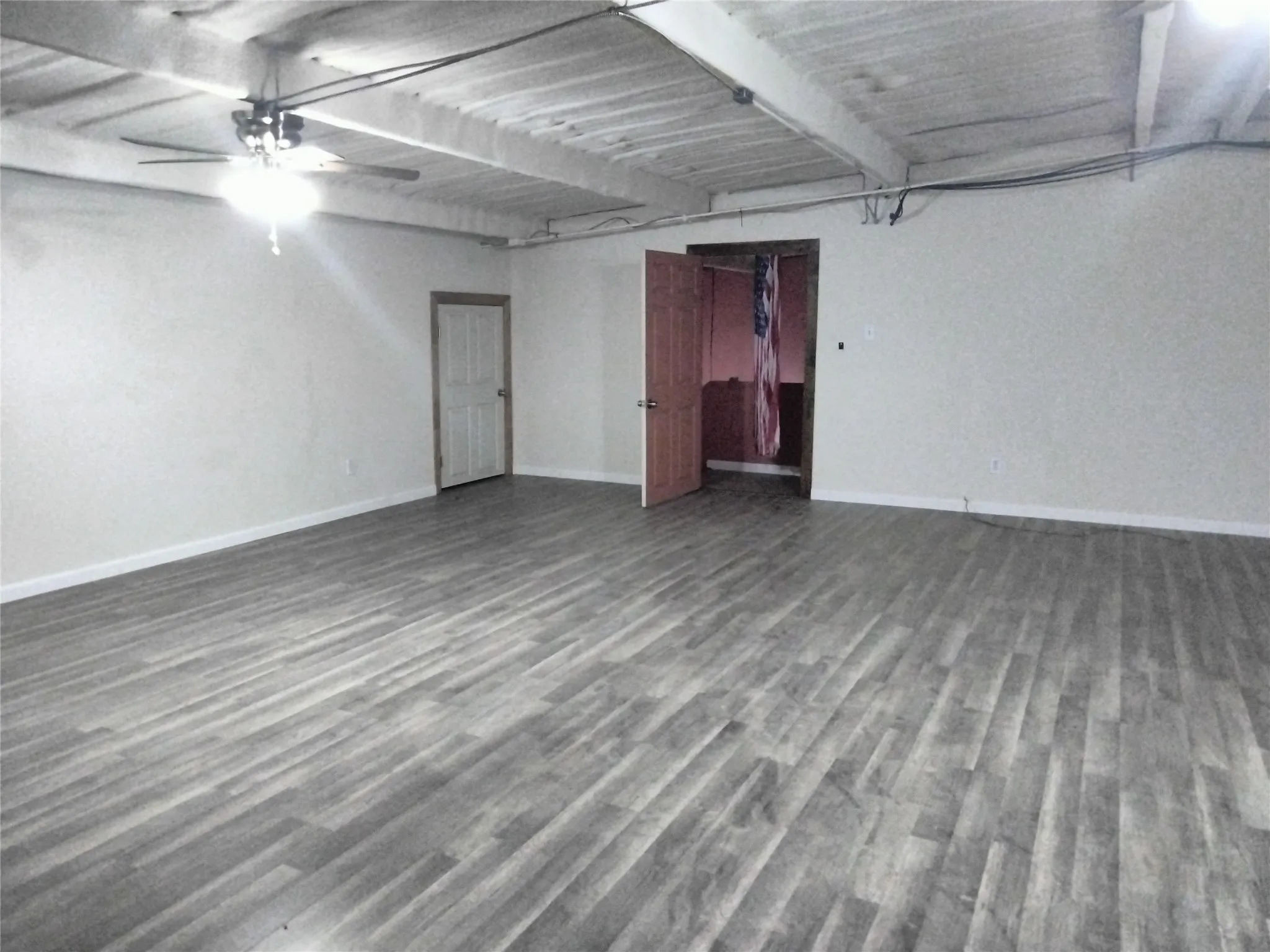 Spacious room featuring wood-finish flooring, a ceiling fan with integrated lighting, and exposed ceiling beams