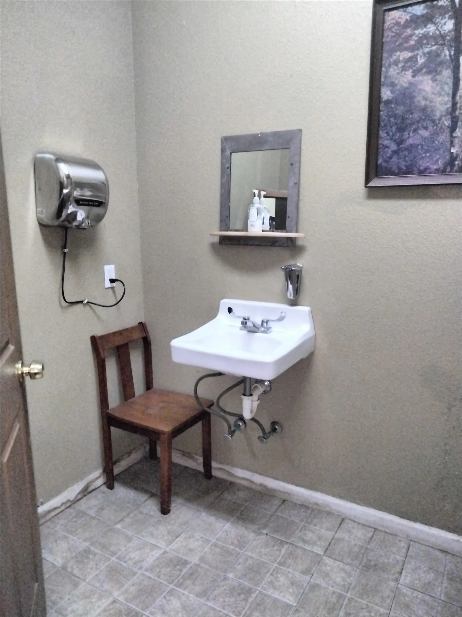 White wall-mounted sink with chrome fixtures, framed mirror with integrated shelf, tiled flooring, and a wall-mounted hand dryer