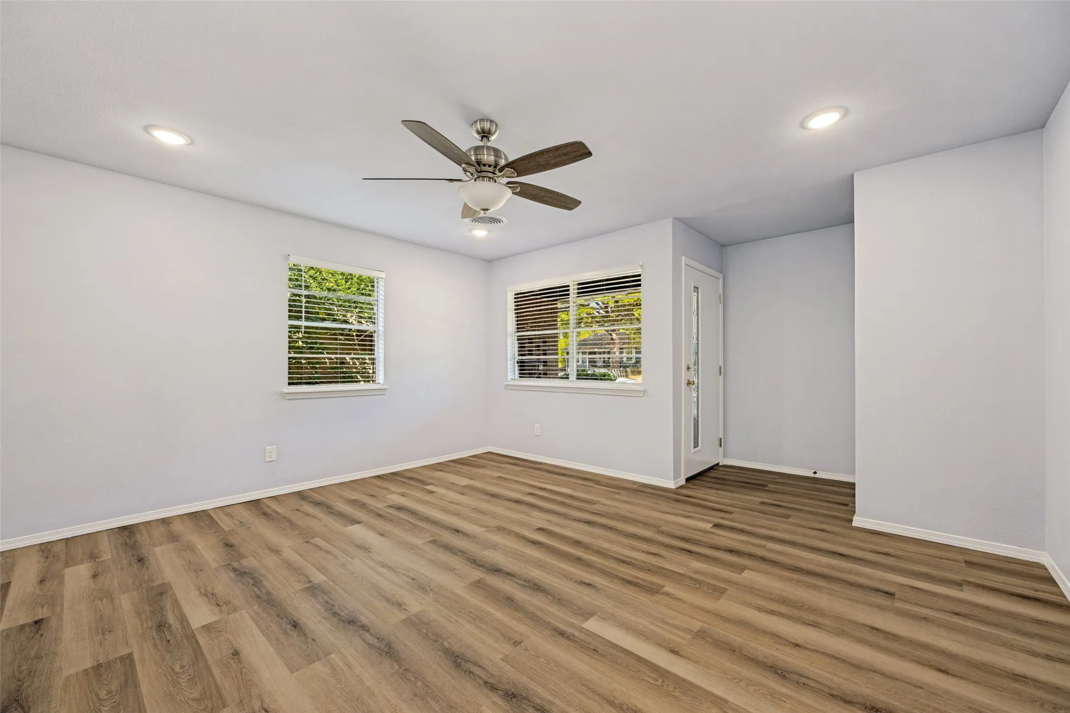 Expansive room featuring wood-finish flooring, two windows with blinds, a ceiling fan, recessed lighting, and a white entry door
