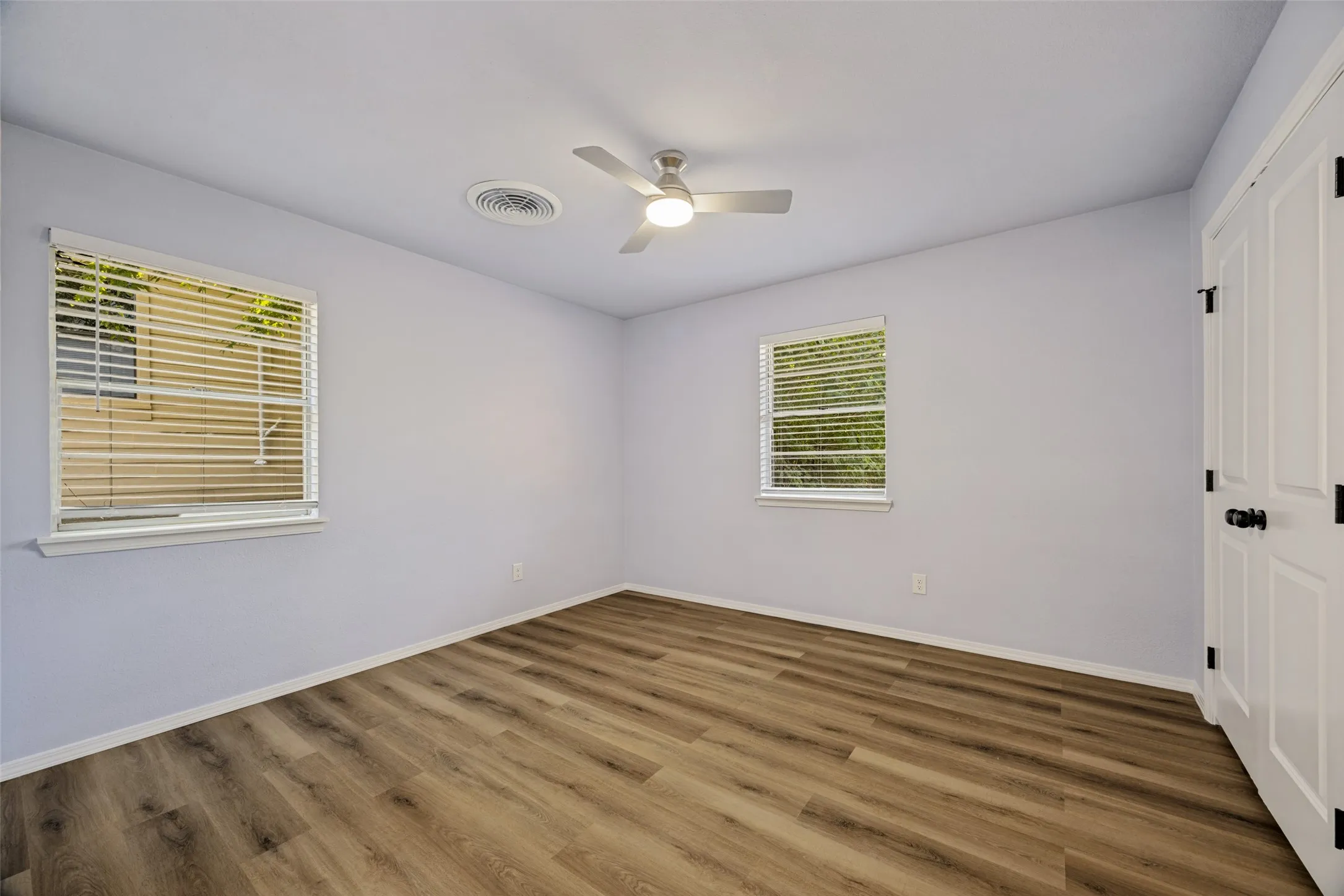 Wood-finish flooring, two windows with blinds, white panel door with black hardware, and a ceiling fan with integrated lighting