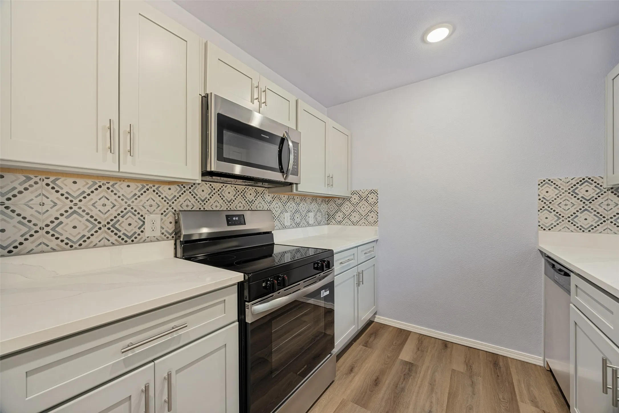 Wood-finish flooring, white countertops, built-in stainless steel microwave, black and stainless steel range, and patterned tile backsplash