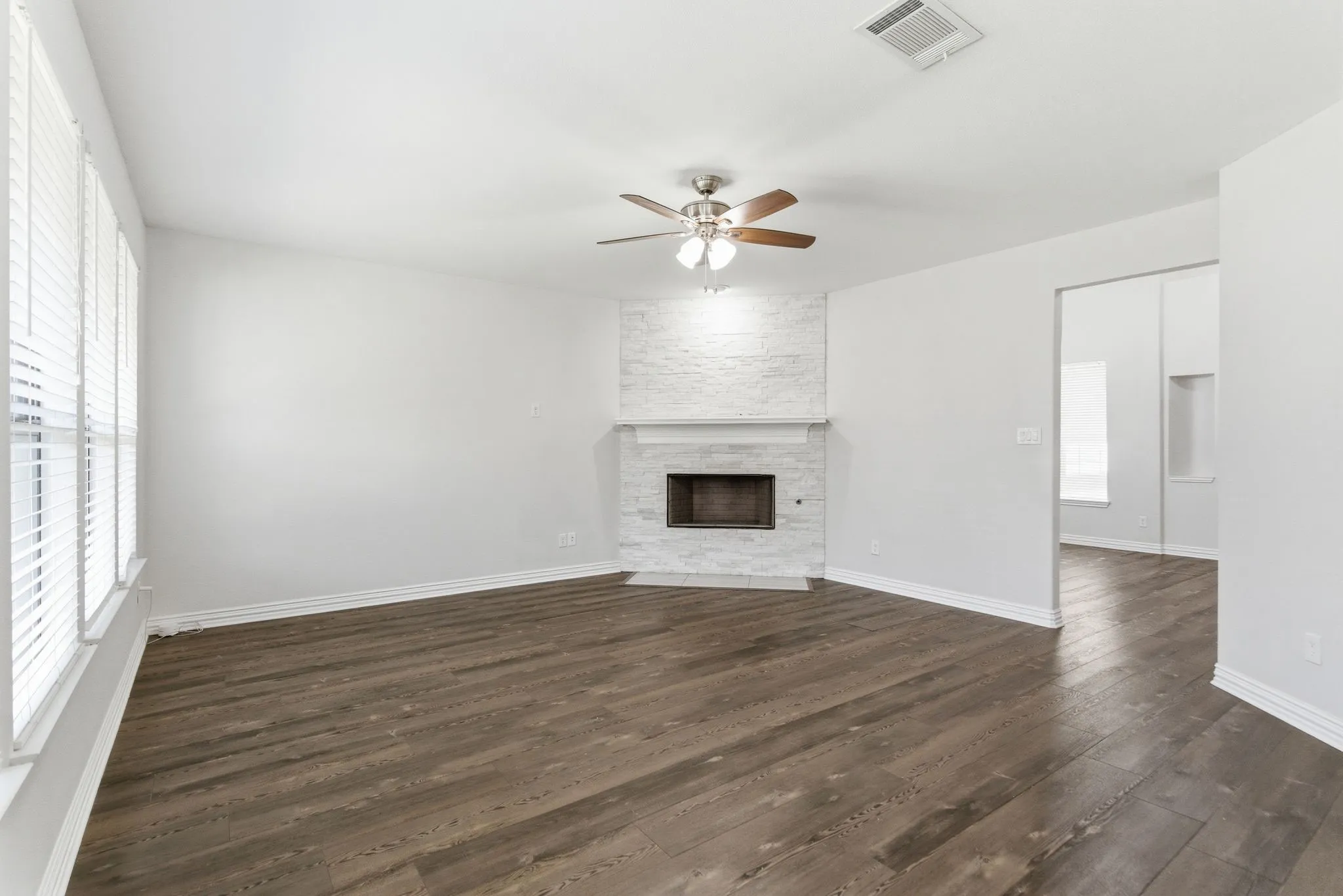 Spacious room featuring wood-finish flooring, a white brick fireplace with a mantel, a ceiling fan, and multiple windows with blinds