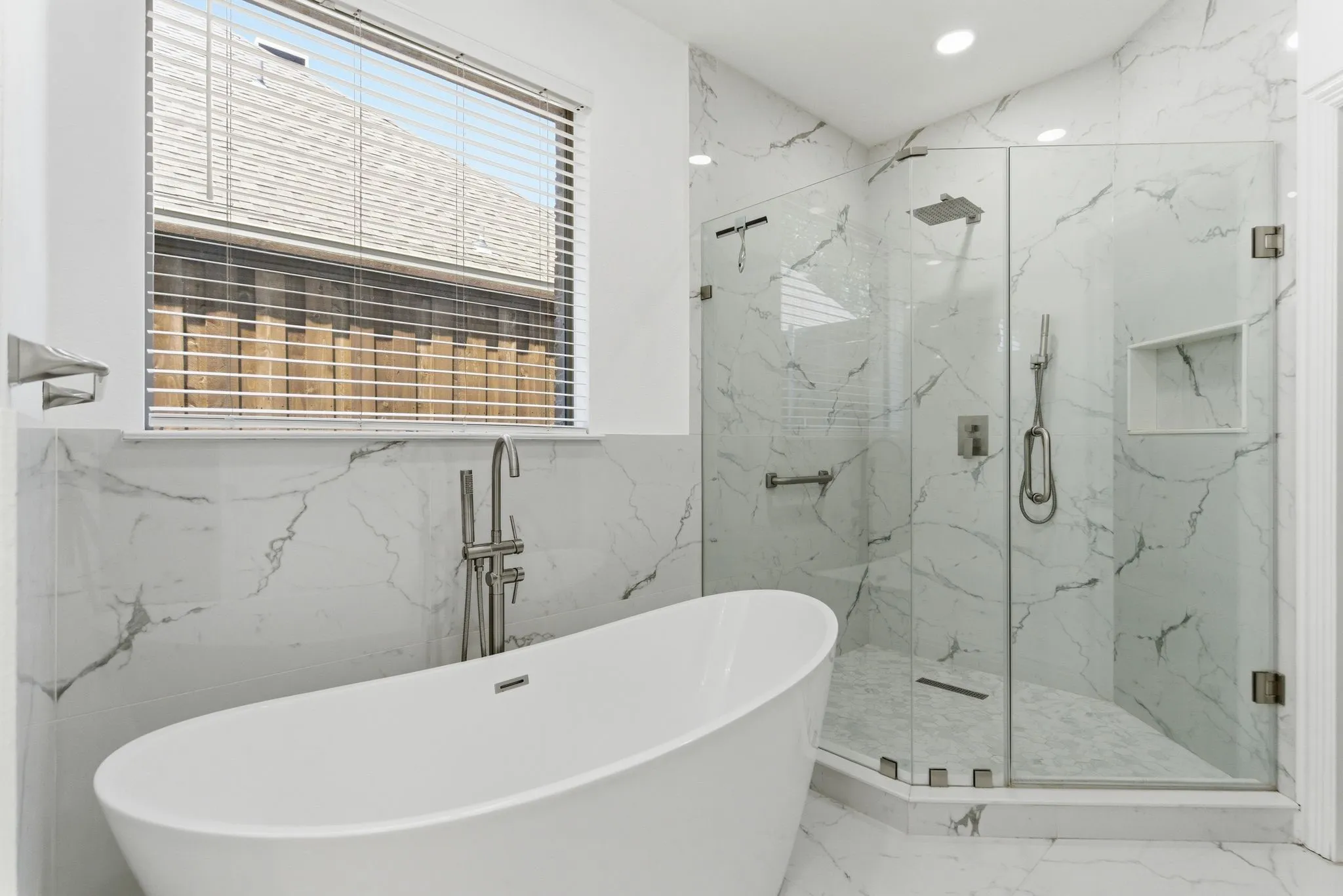 Freestanding soaking tub with brushed nickel floor-mounted filler, glass-enclosed shower, shower niche, and white marble-look wall and floor tile