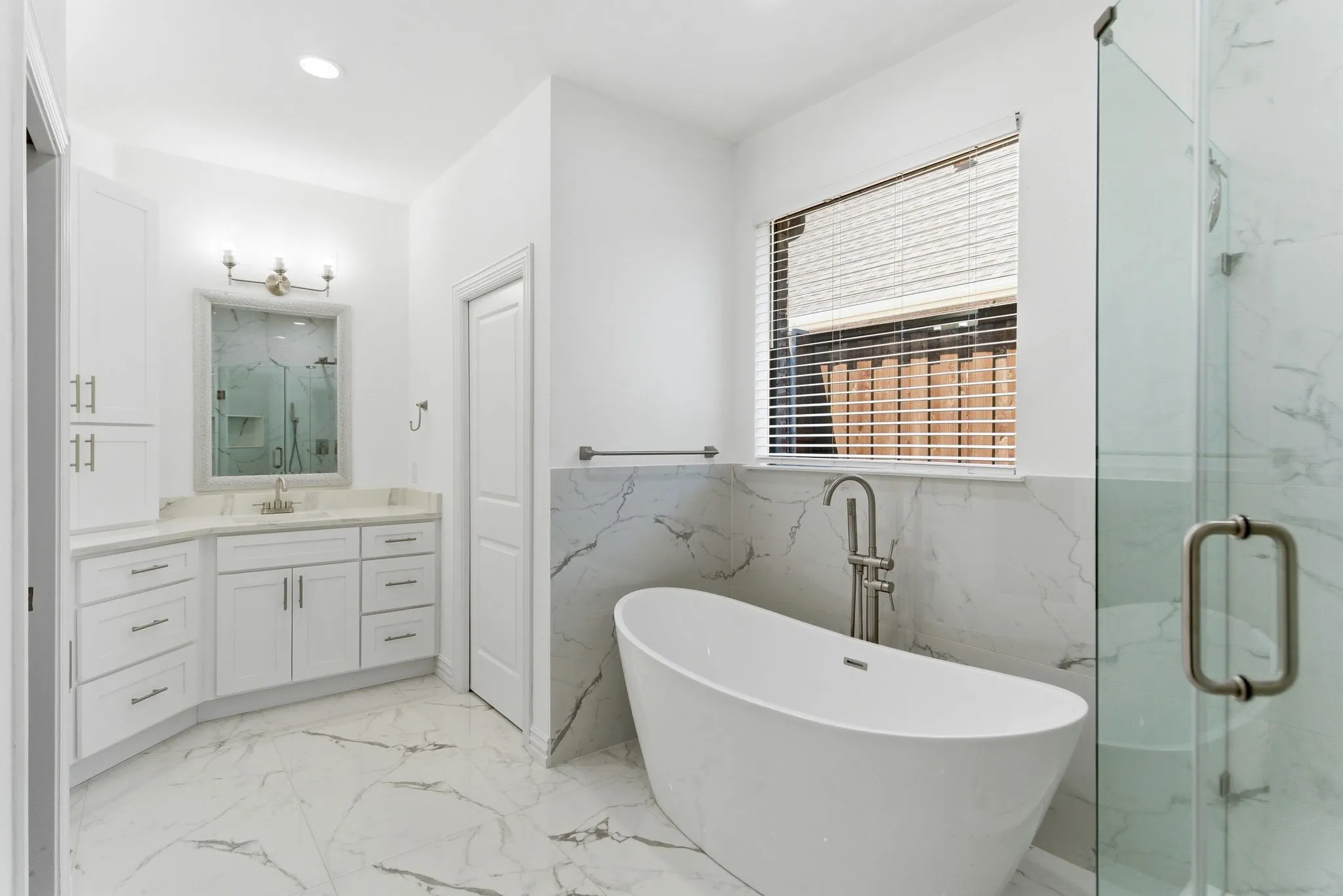 Freestanding soaking tub with floor-mounted faucet, glass-enclosed shower, dual vanity with undermount sink, white cabinetry with silver hardware, and patterned floor tiling