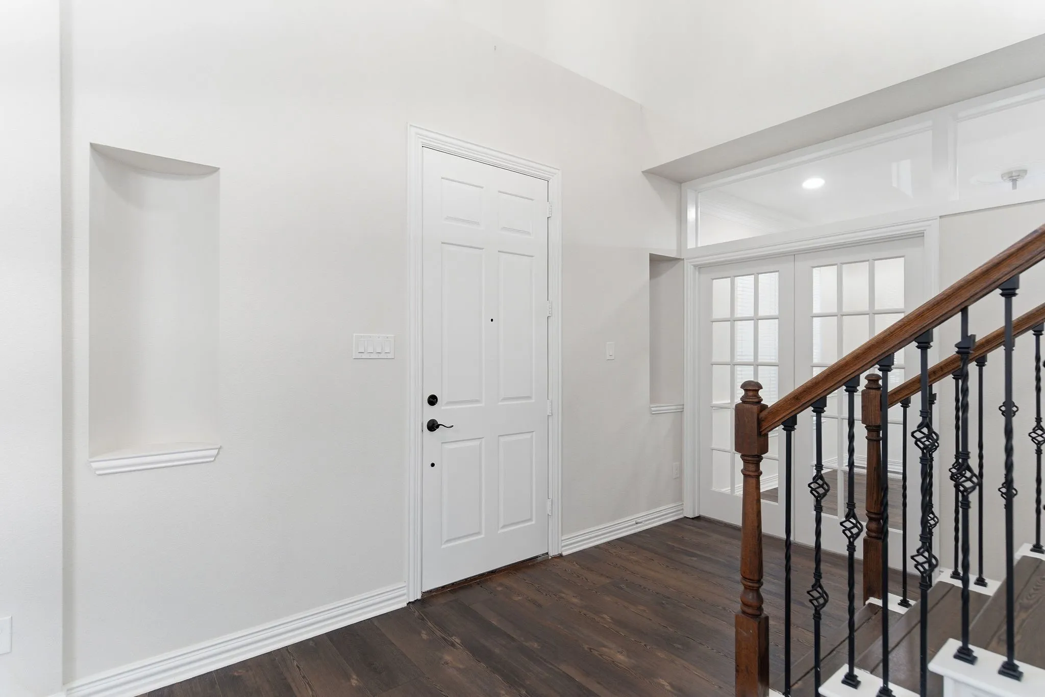Wood-finish flooring, staircase with wrought iron balusters and wood handrail, paneled entry door, recessed wall niche, and double French doors with transom
