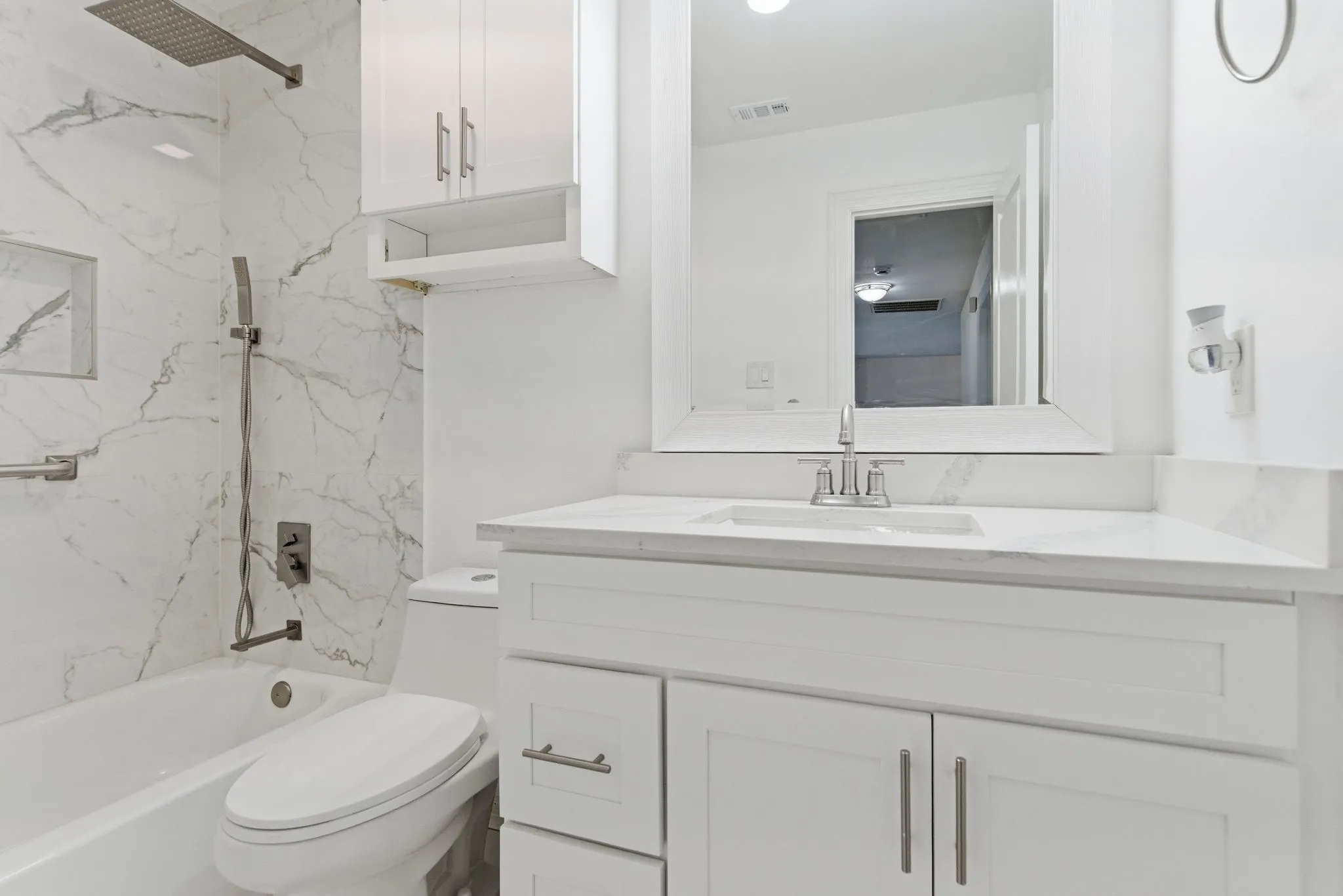 Bathtub with marble-patterned tile surround, rainfall showerhead, white vanity with integrated sink, and upper cabinetry