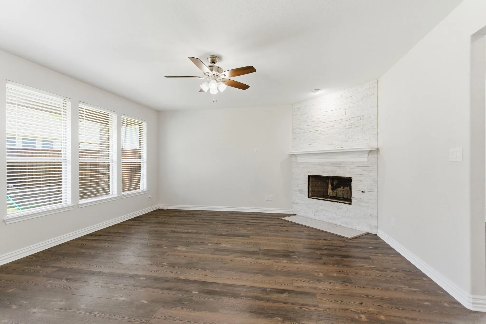 Wood-finish flooring, white walls, multiple windows with blinds, a ceiling fan, and a fireplace with a white brick surround