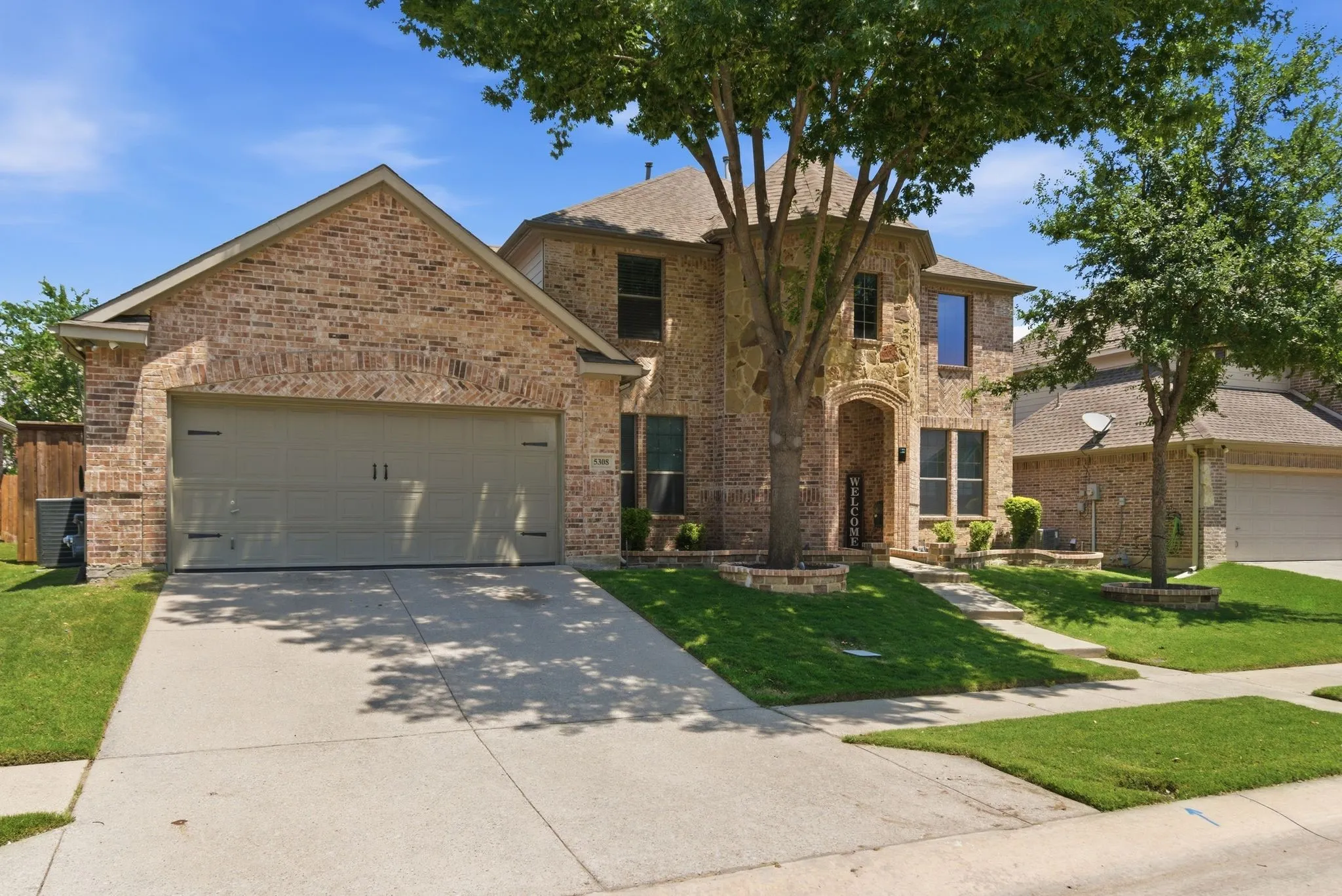 Brick and stone facade, arched entryway, attached garage, well-maintained landscaping, and mature trees