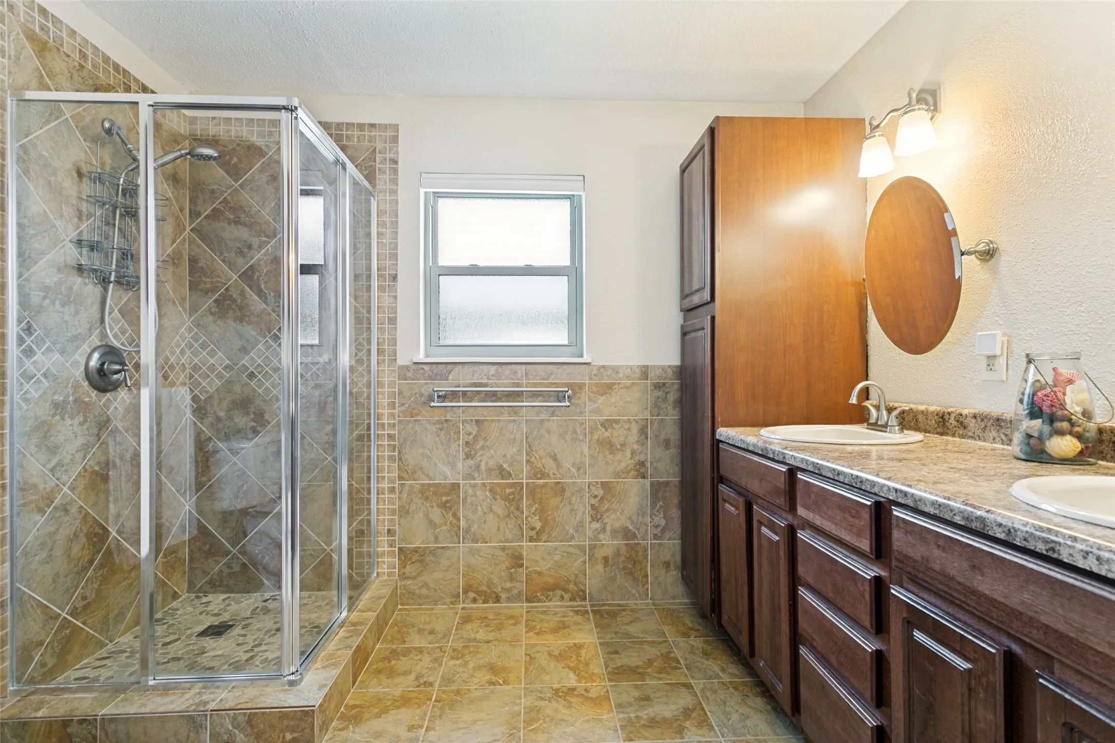 Dual vanity with individual sinks, separate shower enclosure with tile surround, built-in tall storage cabinetry, and window with privacy glass