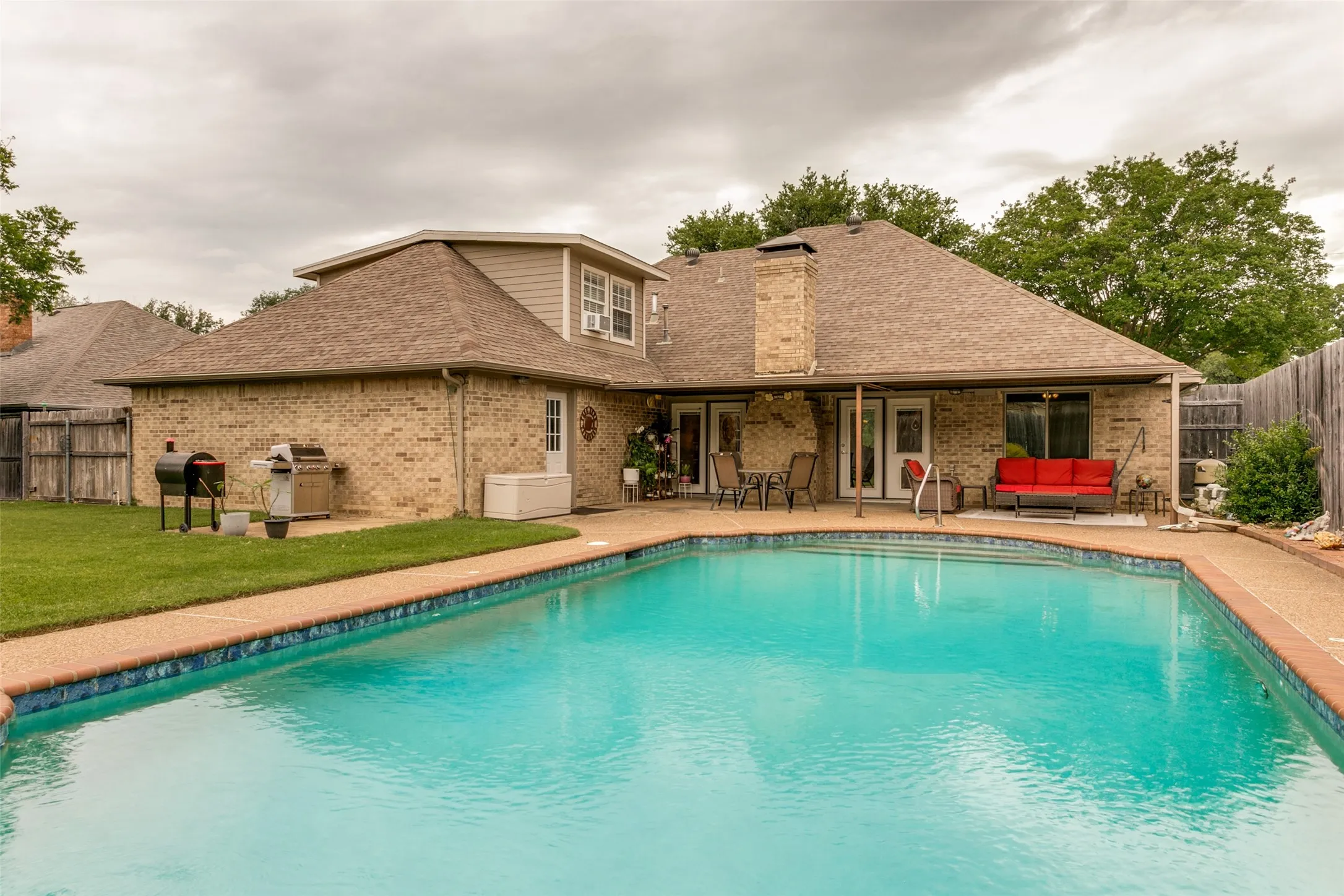 Swimming pool with integrated steps, brick exterior, covered patio, privacy fencing, and a manicured lawn
