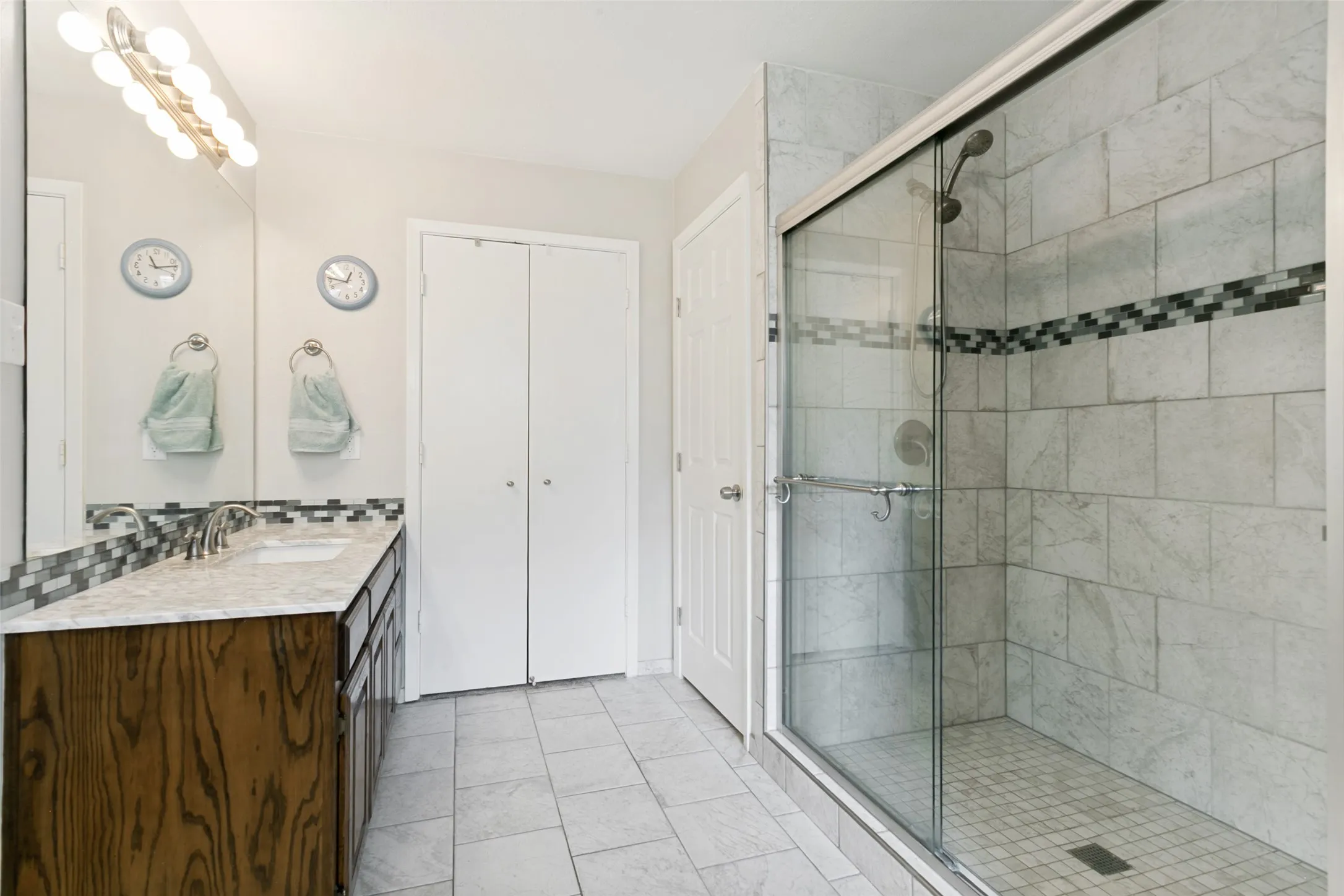 Spacious bathroom featuring oak vaniies with a single sink, an expansive mirror, and a tiled shower with a glass enclosure