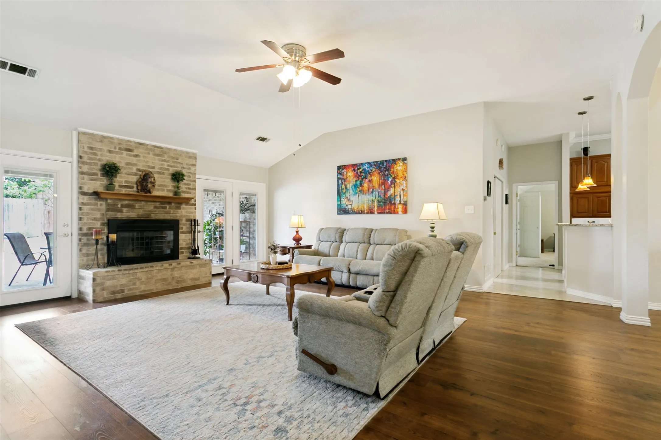 Vaulted ceiling with a ceiling fan, brick fireplace with a wood mantel, wood-finish flooring, and multiple glass-paneled doors