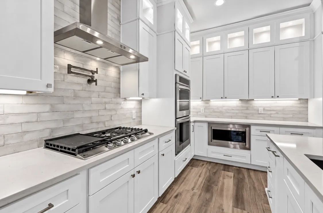 White shaker cabinetry with illuminated upper display cabinets, stainless steel appliances, light-toned brick-patterned backsplash, light-toned countertops, and wood-finish flooring