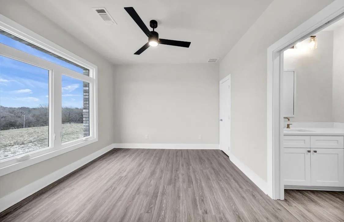 Spacious room featuring wood-finish flooring, large windows, an overhead ceiling fan, and access to a bathroom with a white vanity and single-basin sink
