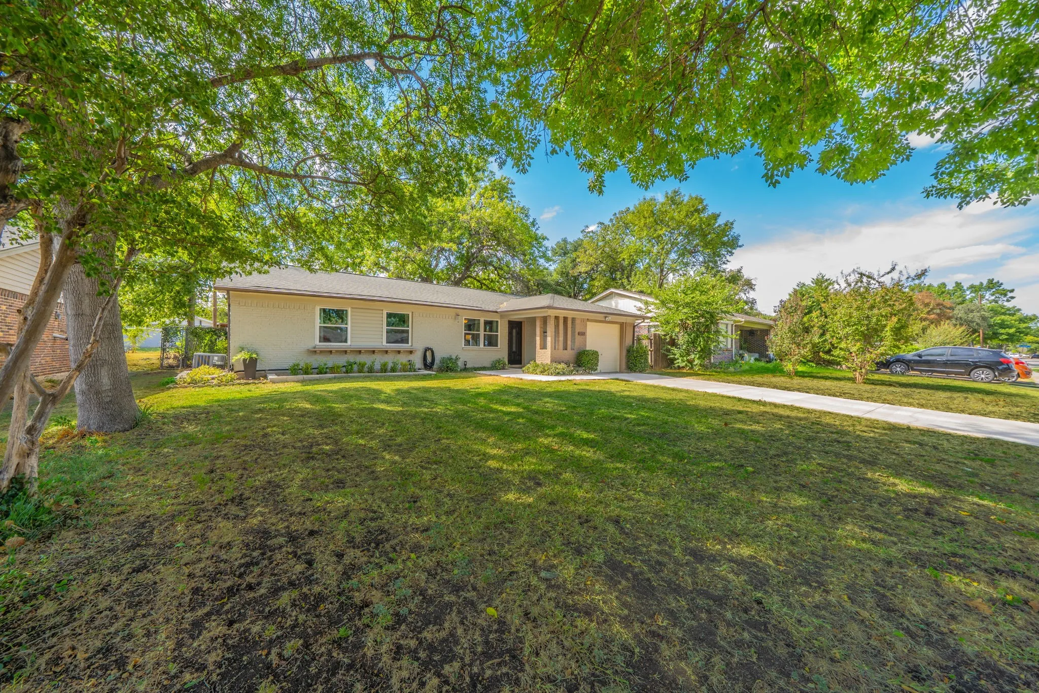 Single-story residence featuring a light-colored exterior, an attached garage, a paved driveway, and mature landscaping