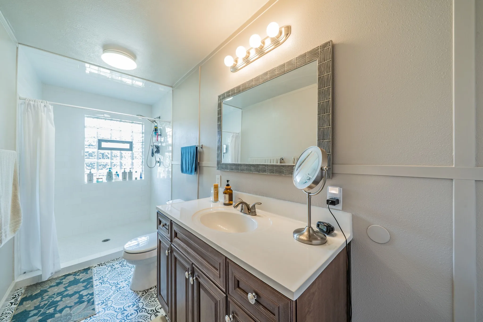 Vanity with a white countertop and wood-finish cabinetry, framed mirror with integrated lighting, shower with a glass block window, and patterned floor tiling