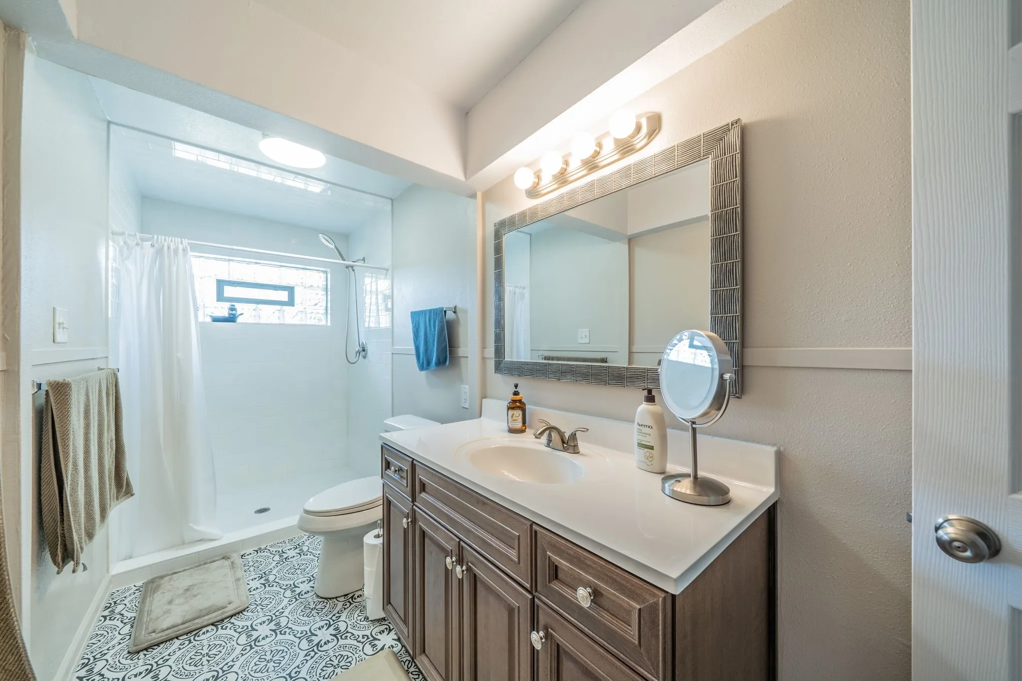 Bathroom featuring a wood-finish vanity with a single basin sink, a framed mirror, an overhead vanity light fixture, a walk-in shower with a glass panel, and patterned floor tile