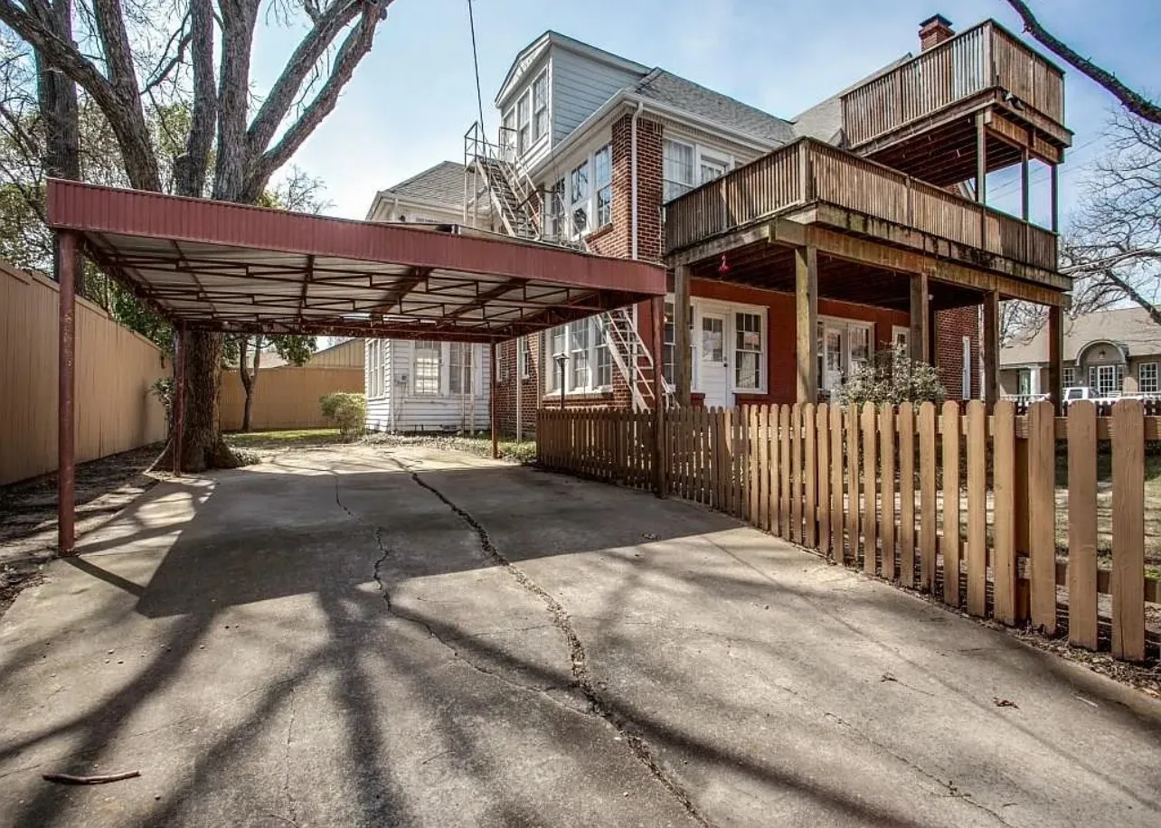 Covered carport, multi-level wooden deck, brick exterior, picket fence, and mature tree
