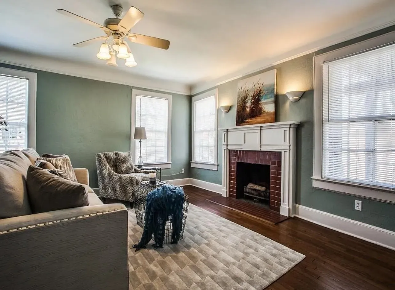 Wood-finish flooring, a brick fireplace with a white mantel, multiple windows, built-in wall sconces, and a ceiling fan with integrated lighting