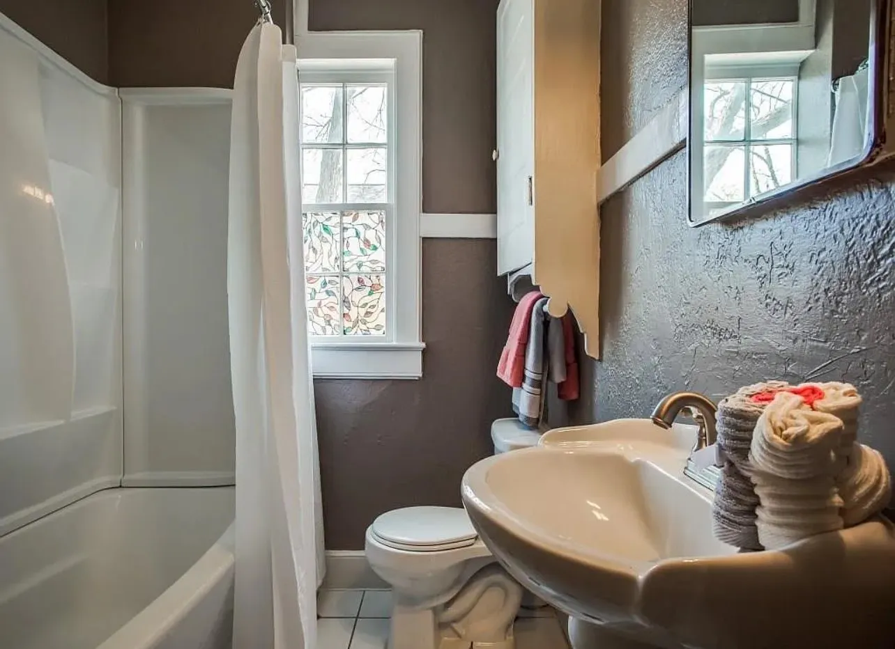 White bathtub with shower, pedestal sink with brass-finish faucet, and a window featuring decorative glass
